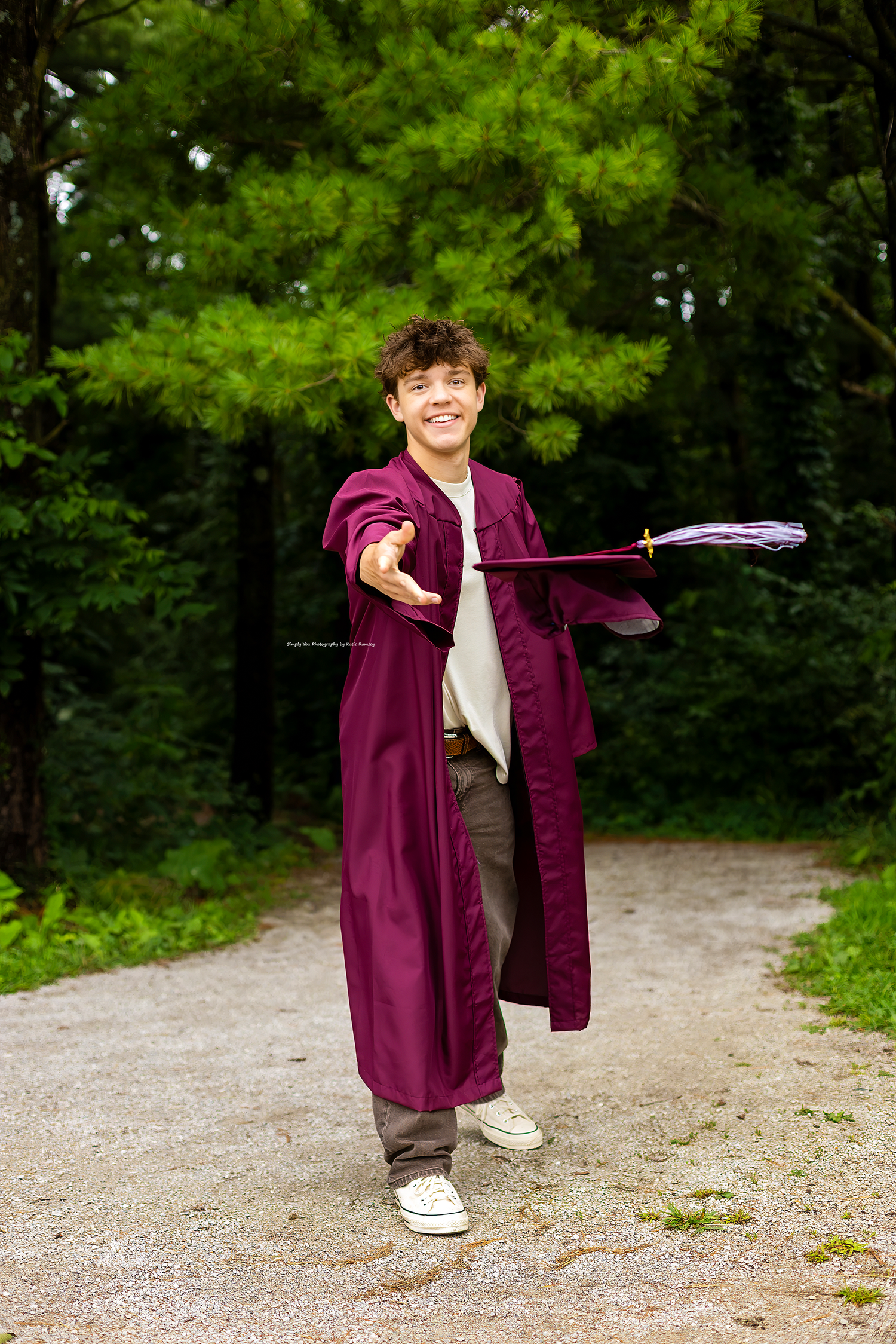 Smiling teen in maroon graduation gown holding a decorated wand on a gravel path surrounded by green trees.