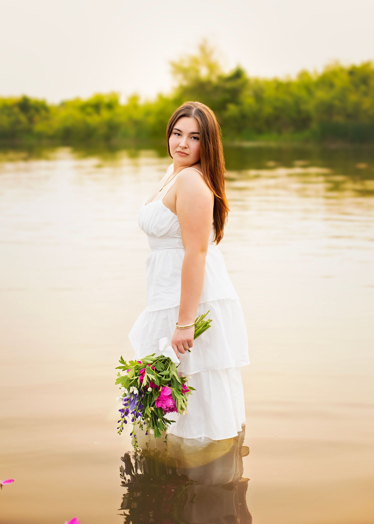 Young woman in white tiered dress holds colorful bouquet while standing in shallow water.
