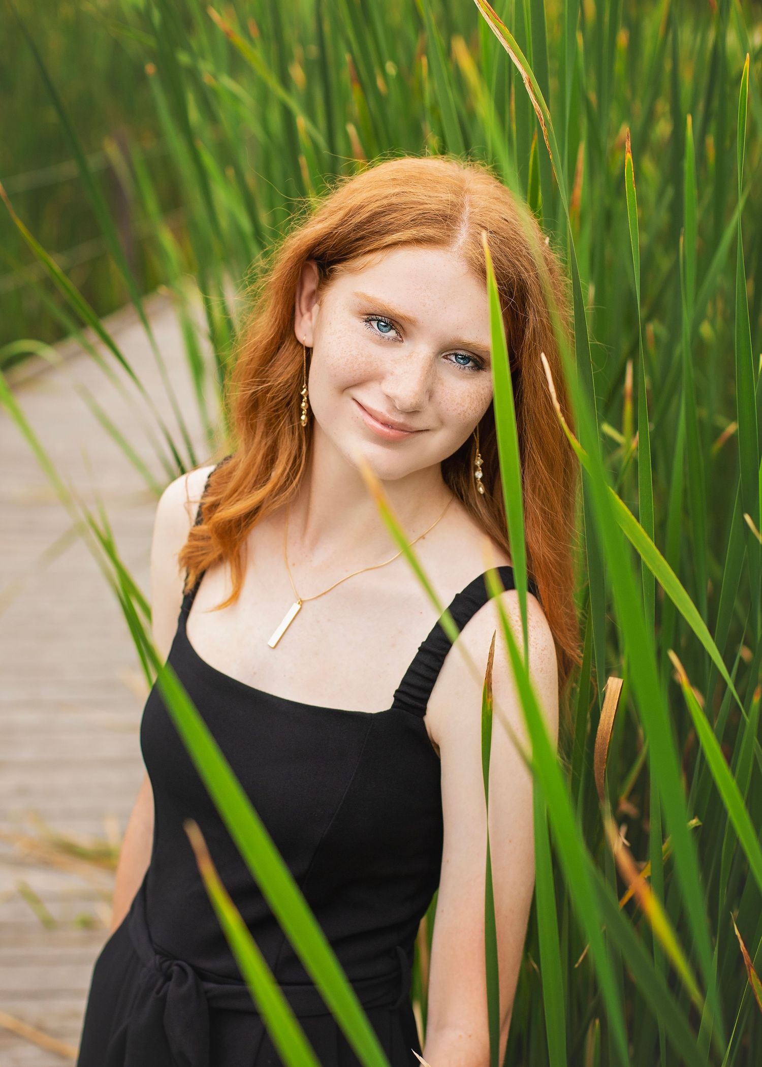 Red-haired teen girl with blue eyes smiles among tall green reeds near a wooden boardwalk.
