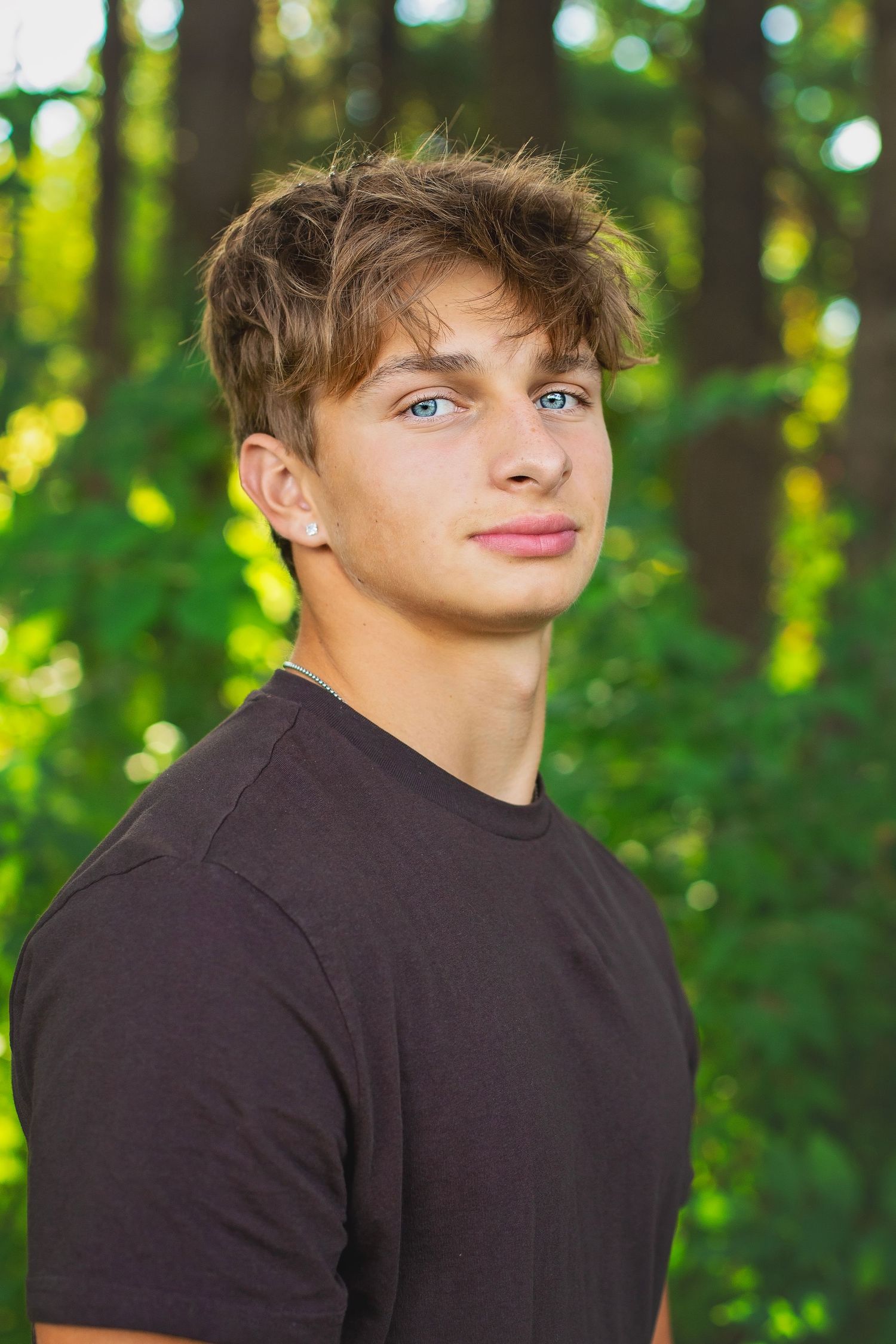 Close-up portrait of a blue-eyed teen boy with tousled blond hair wearing a dark t-shirt in a forest.