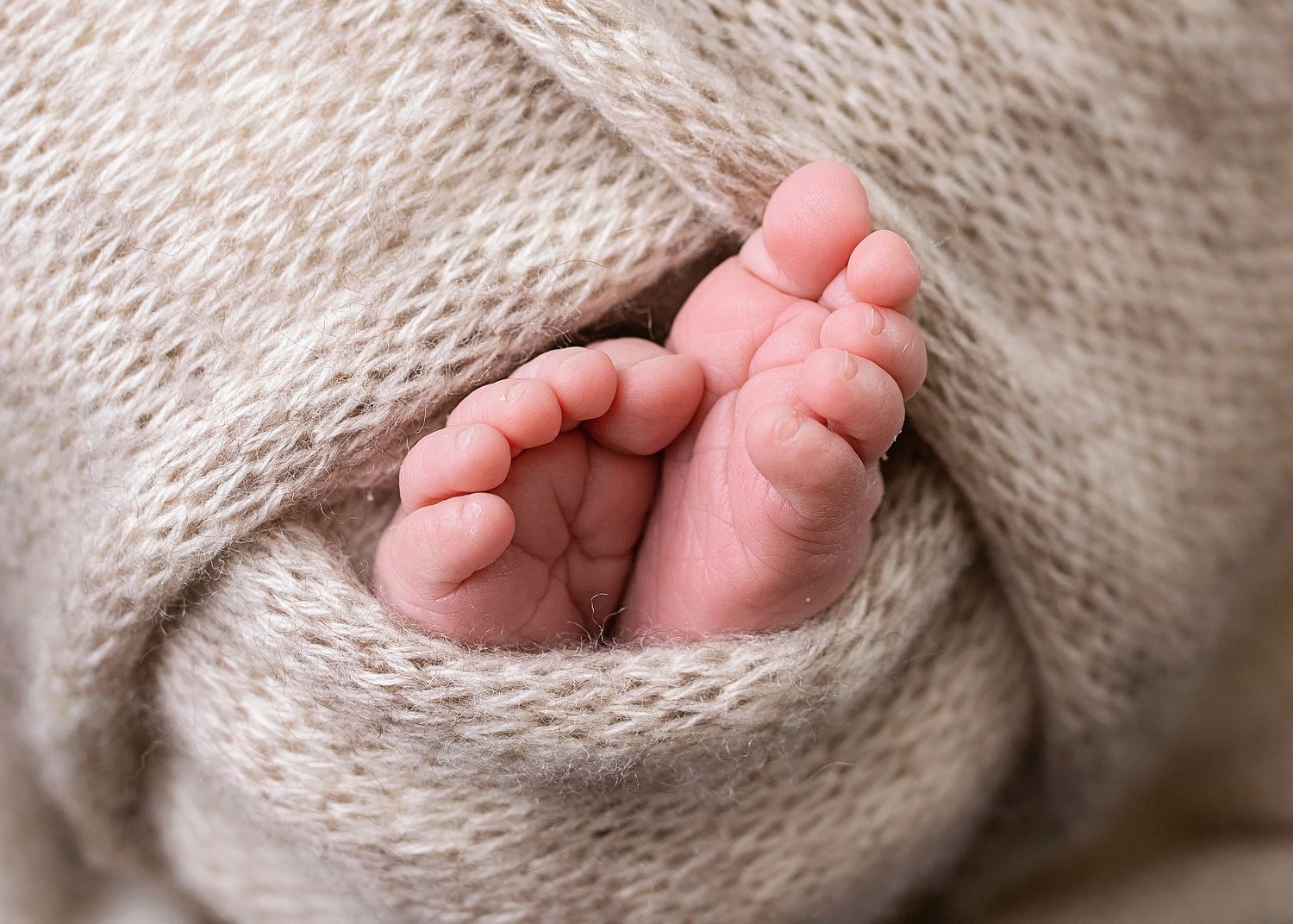 Close-up of tiny newborn baby feet peeking out from a soft beige knit swaddle wrap.