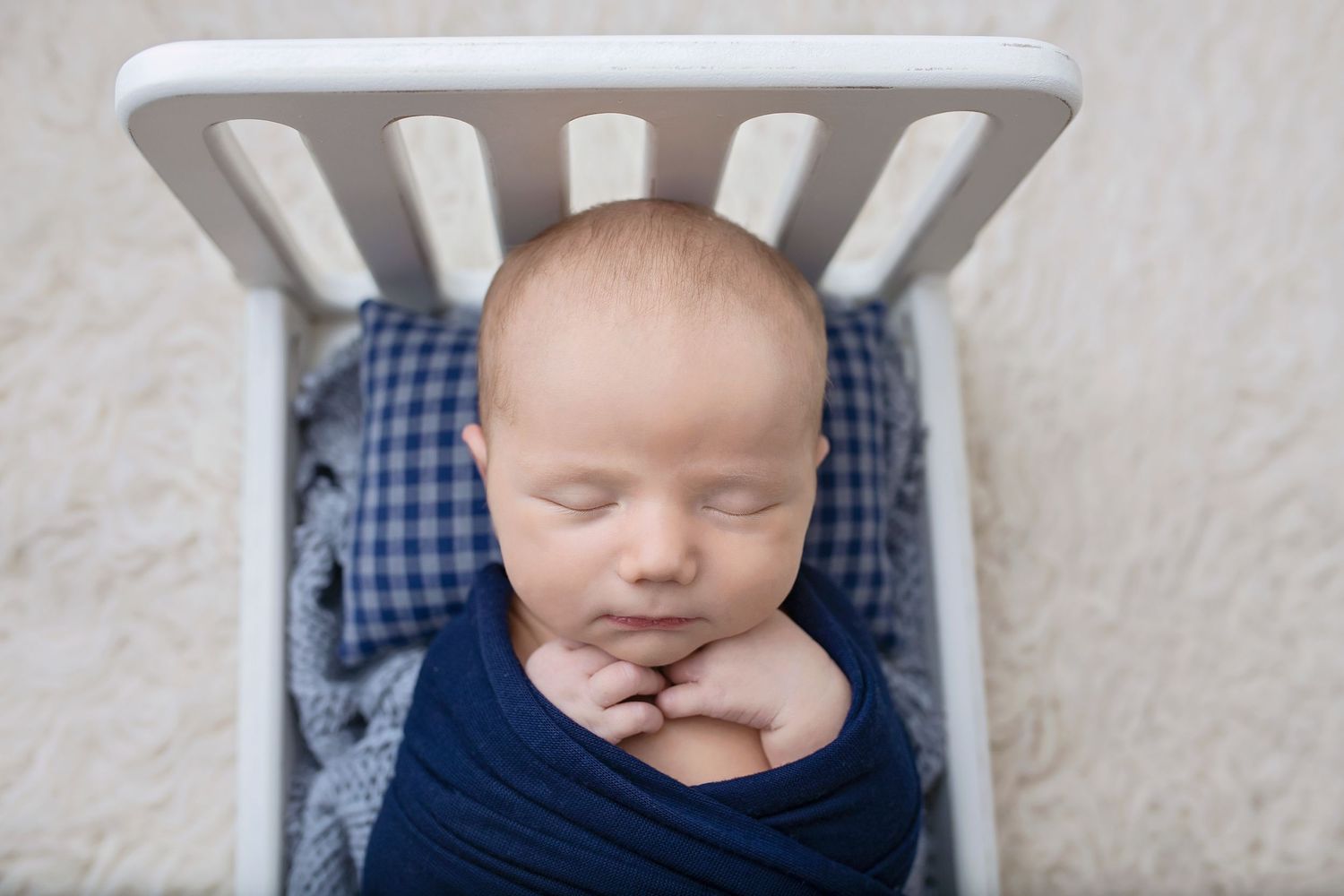 Peacefully sleeping newborn baby swaddled in navy blue wrap in a miniature white crib.