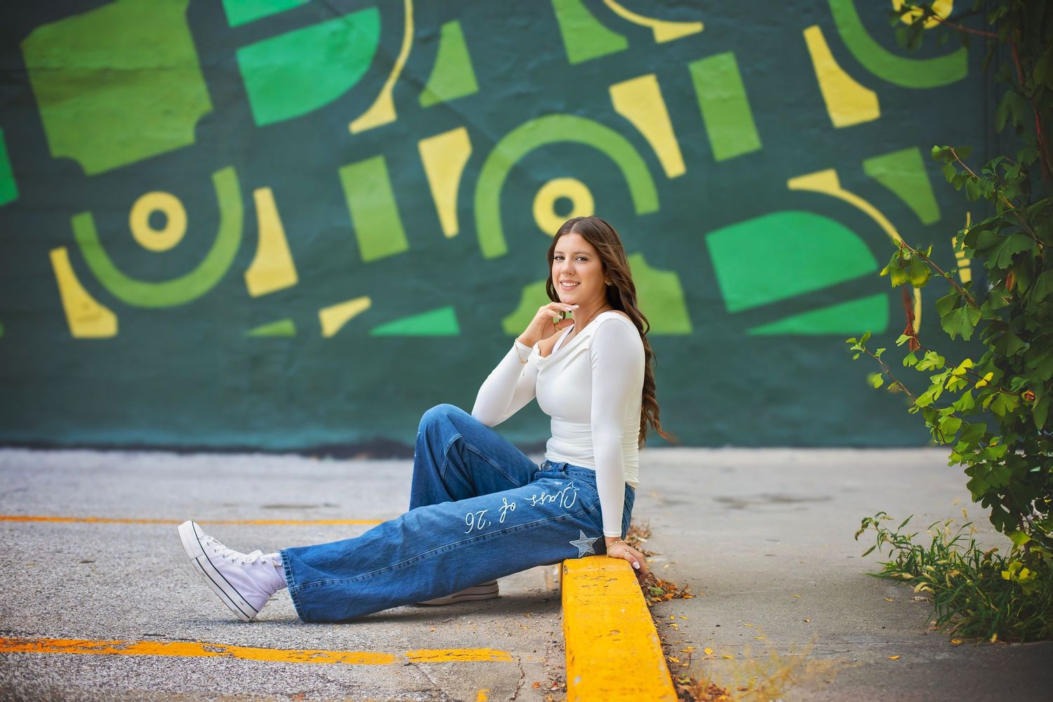 Teen girl in white top and wide-leg jeans sits relaxed before bold green geometric graffiti mural.
