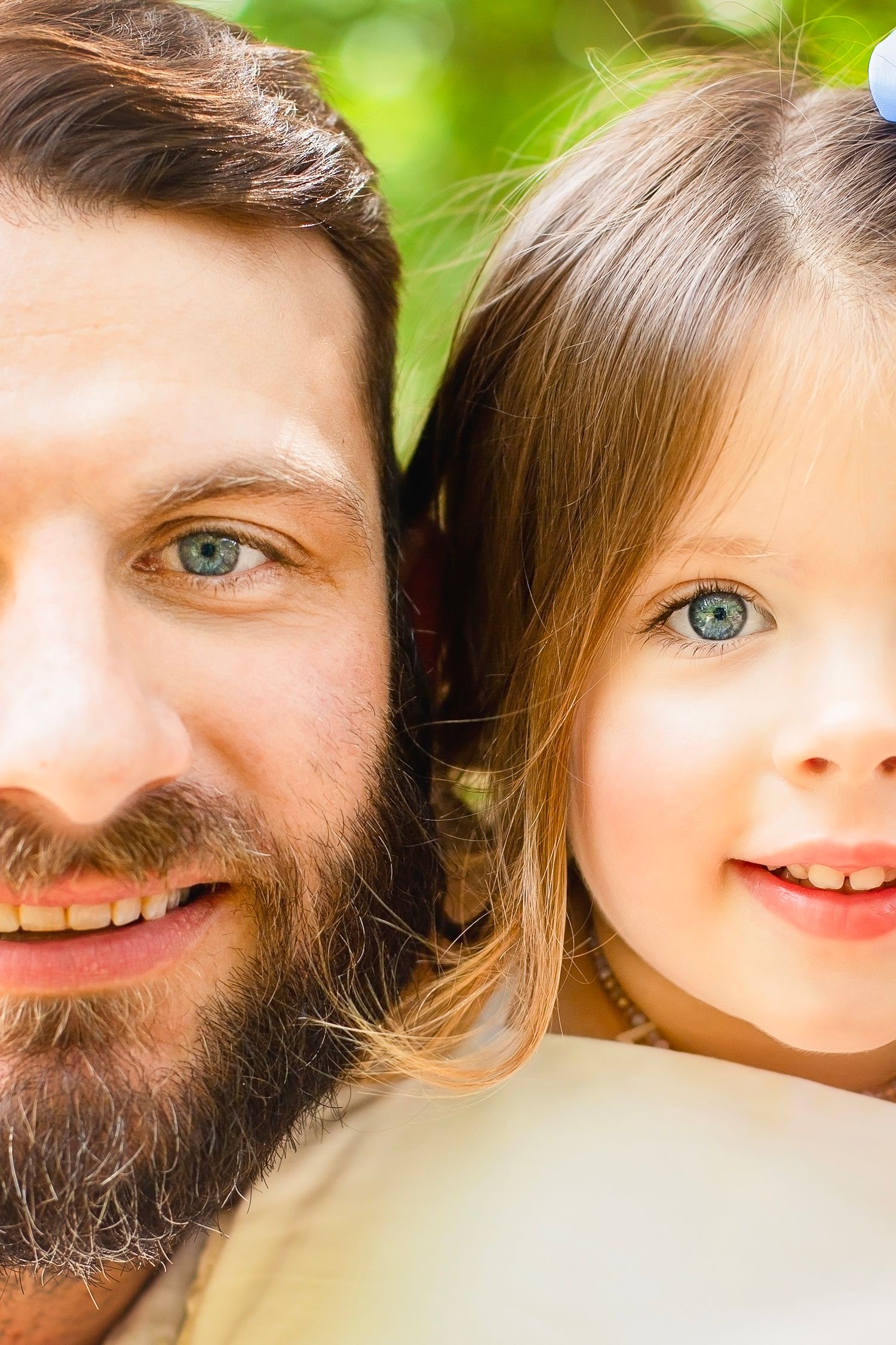 Close-up portrait of bearded father and young daughter sharing matching blue eyes and bright smiles outdoors.