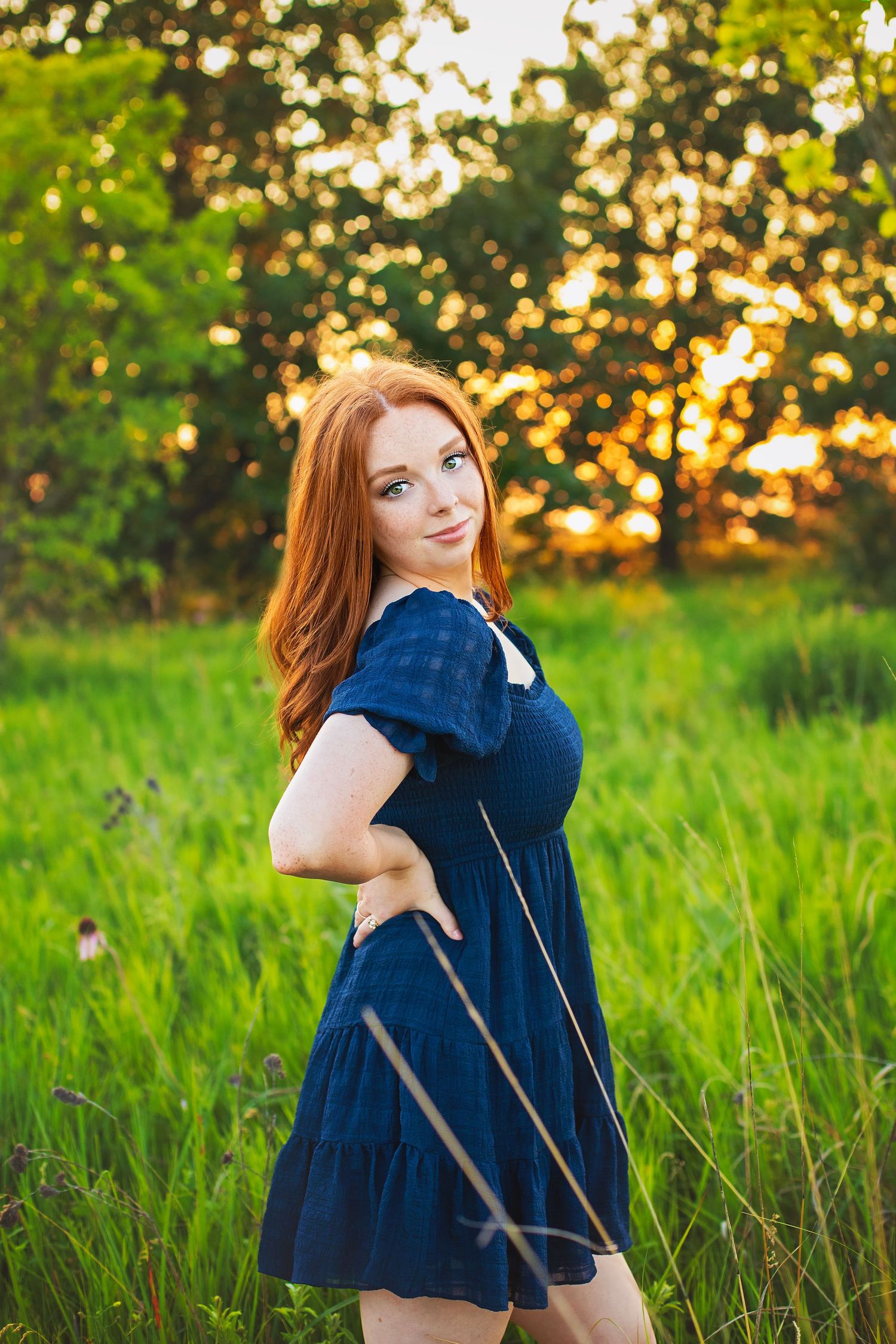 Young woman with red hair in navy dress poses in golden sunset light among lush green meadow grass.