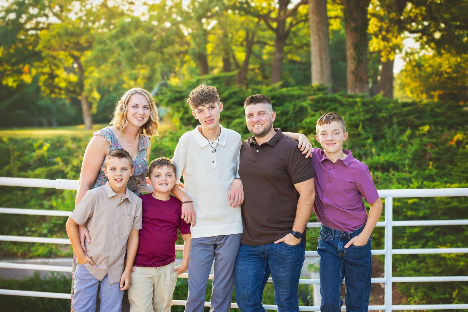 Family of six with a mother and five boys smiling together outdoors in golden sunlight near a white fence.