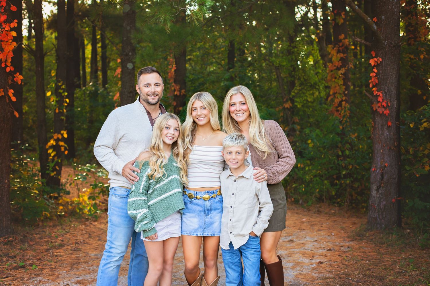 Family of five smiling together on a wooded path with colorful autumn foliage.