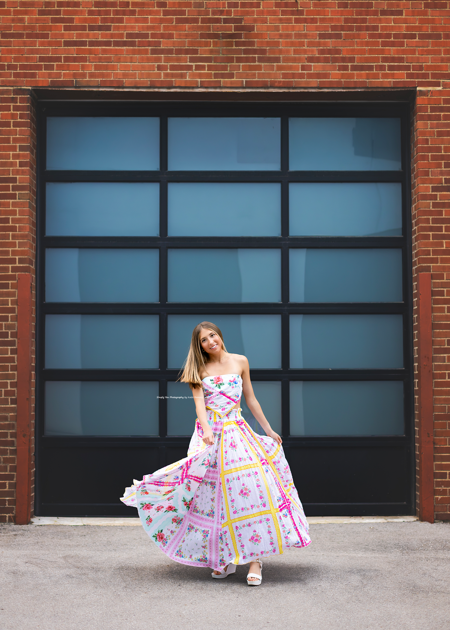 Woman twirling in a colorful floral maxi dress in front of a brick wall with large industrial windows.