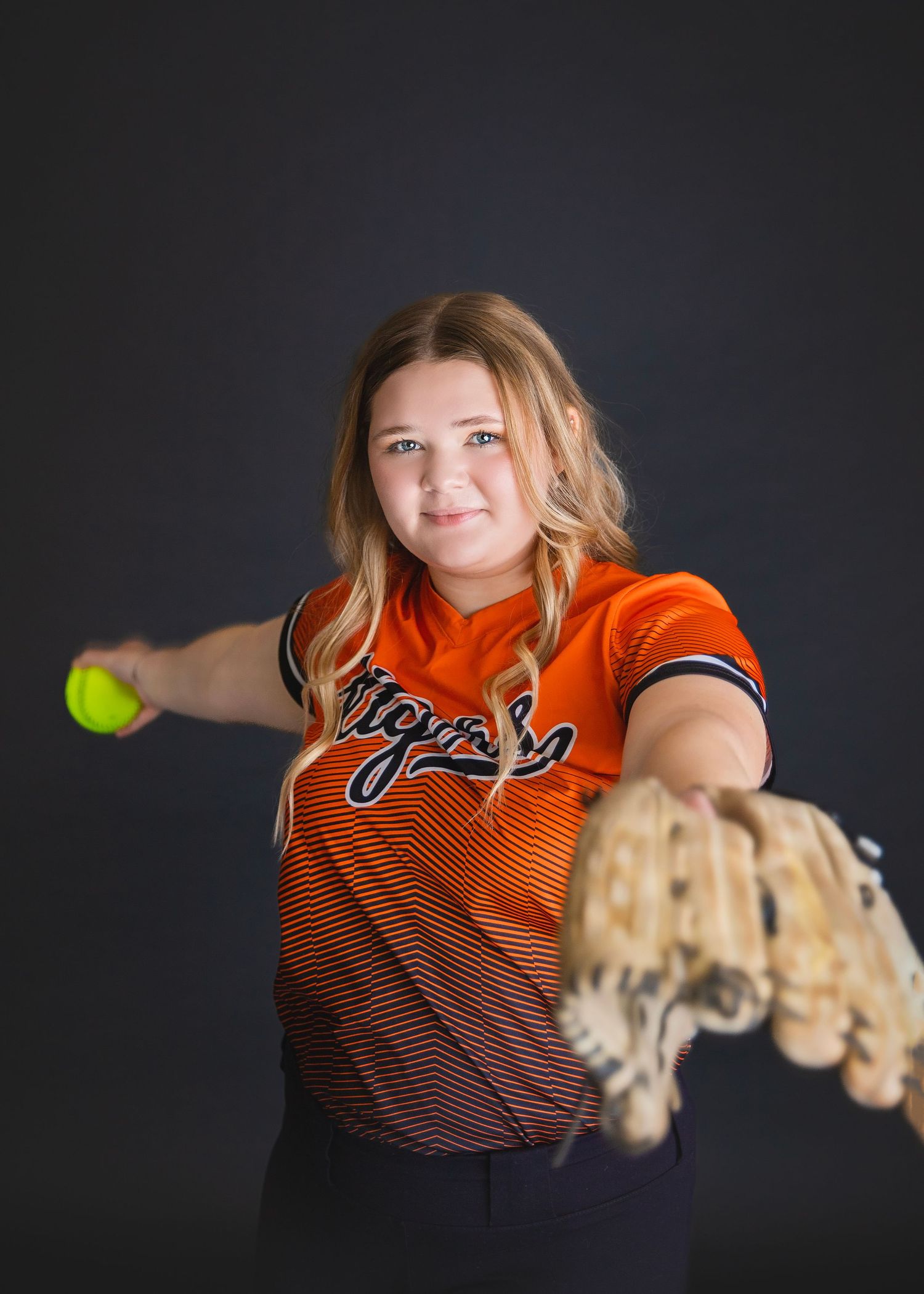 Teen girl in Tigers softball jersey winds up to pitch while holding glove.
