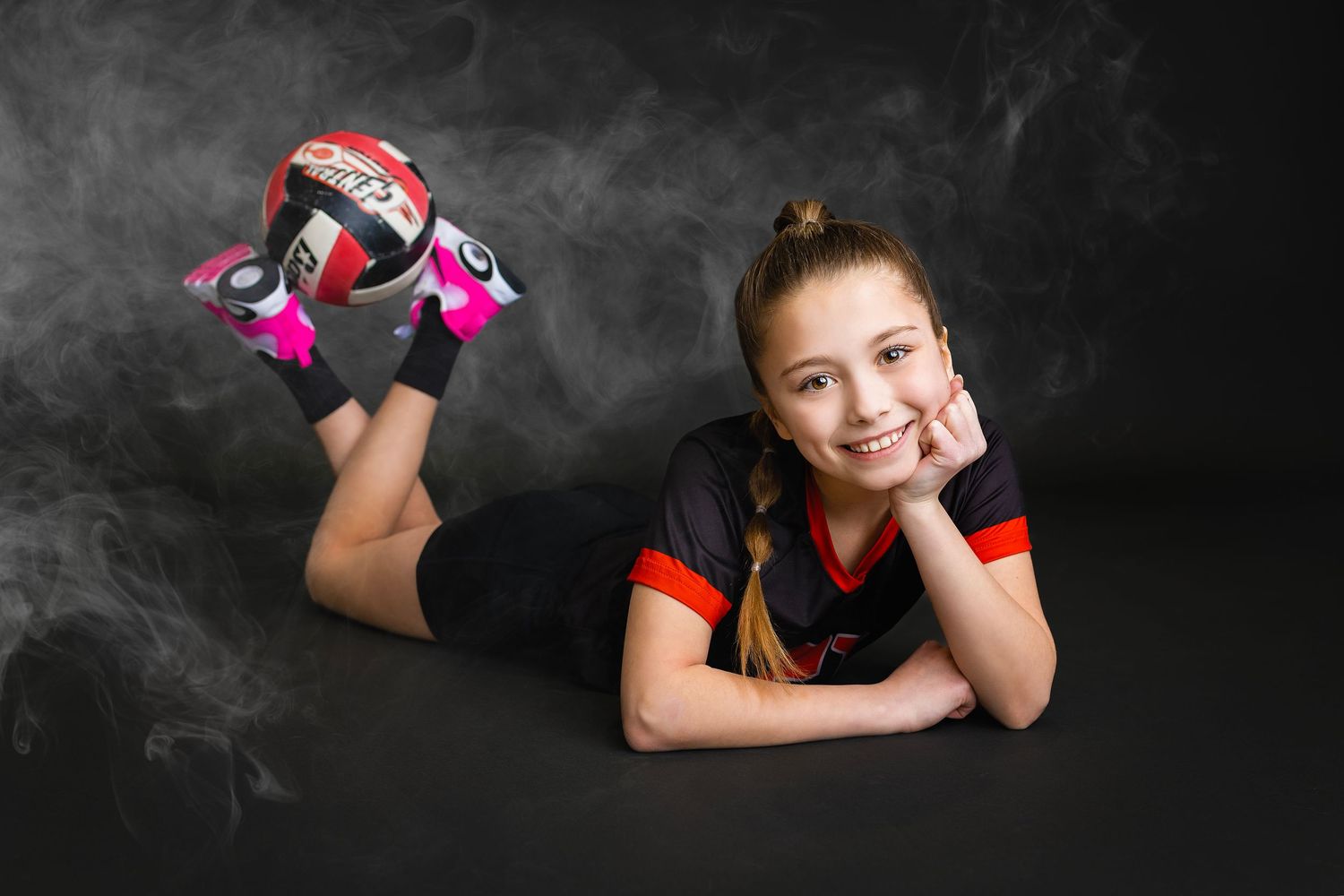 Young girl in black and red volleyball uniform lies on floor smiling with pink and red volleyball shoes raised up.