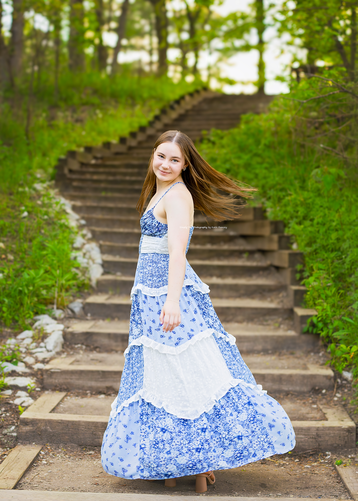 Young girl twirling in blue floral maxi dress on outdoor stone steps surrounded by green trees.