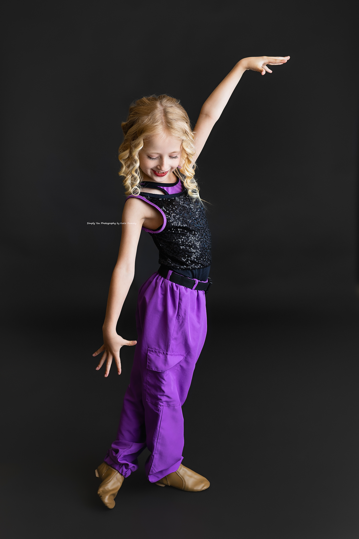 Young blonde girl in purple harem pants and black sequin top strikes a dance pose with arm raised.