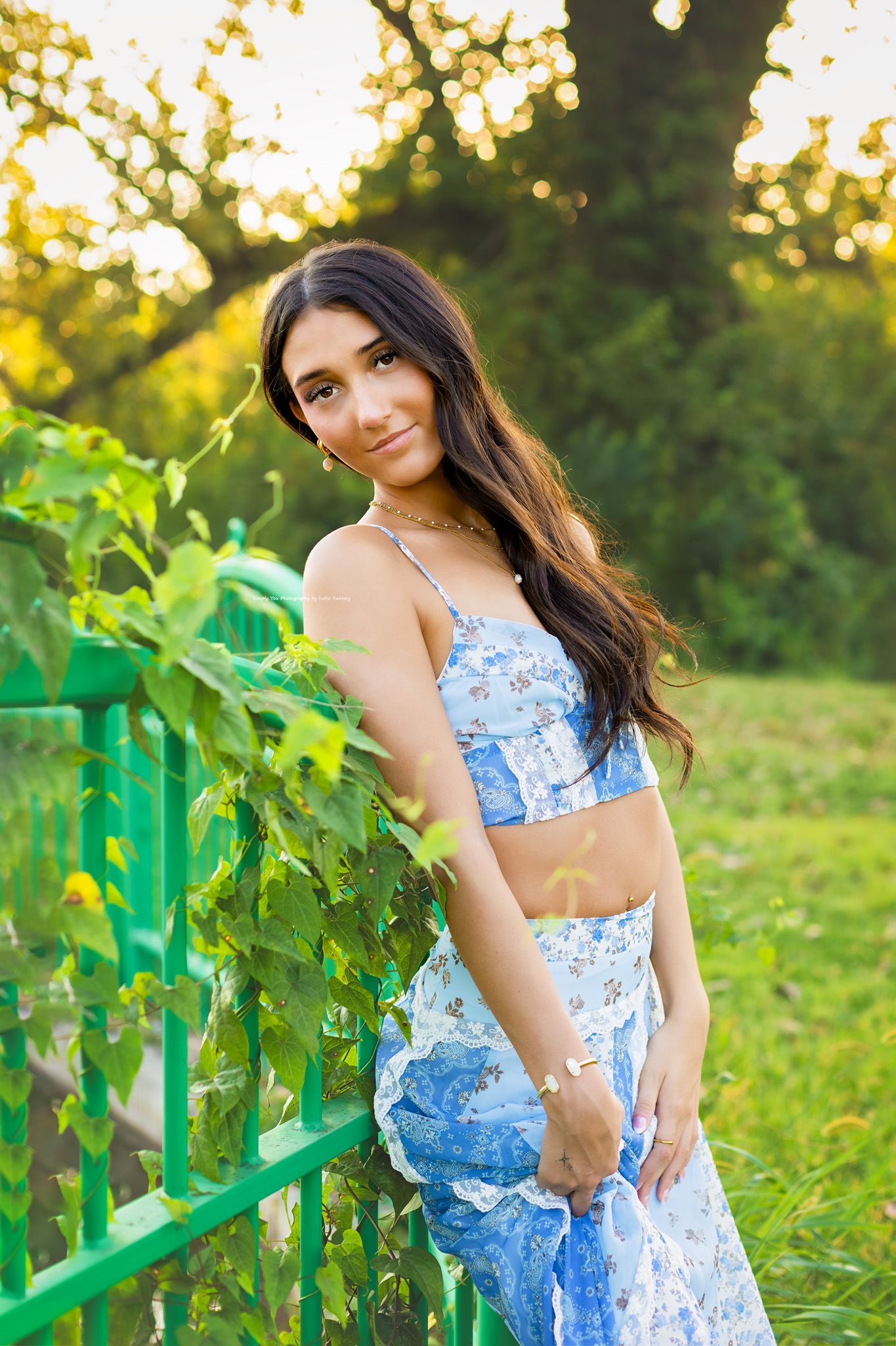Young woman in blue floral two-piece outfit leaning on green fence surrounded by lush greenery in golden hour sunlight.