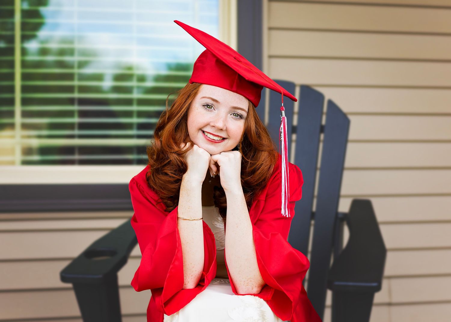 Smiling young woman in red graduation cap and gown sitting in a chair outdoors on a porch.