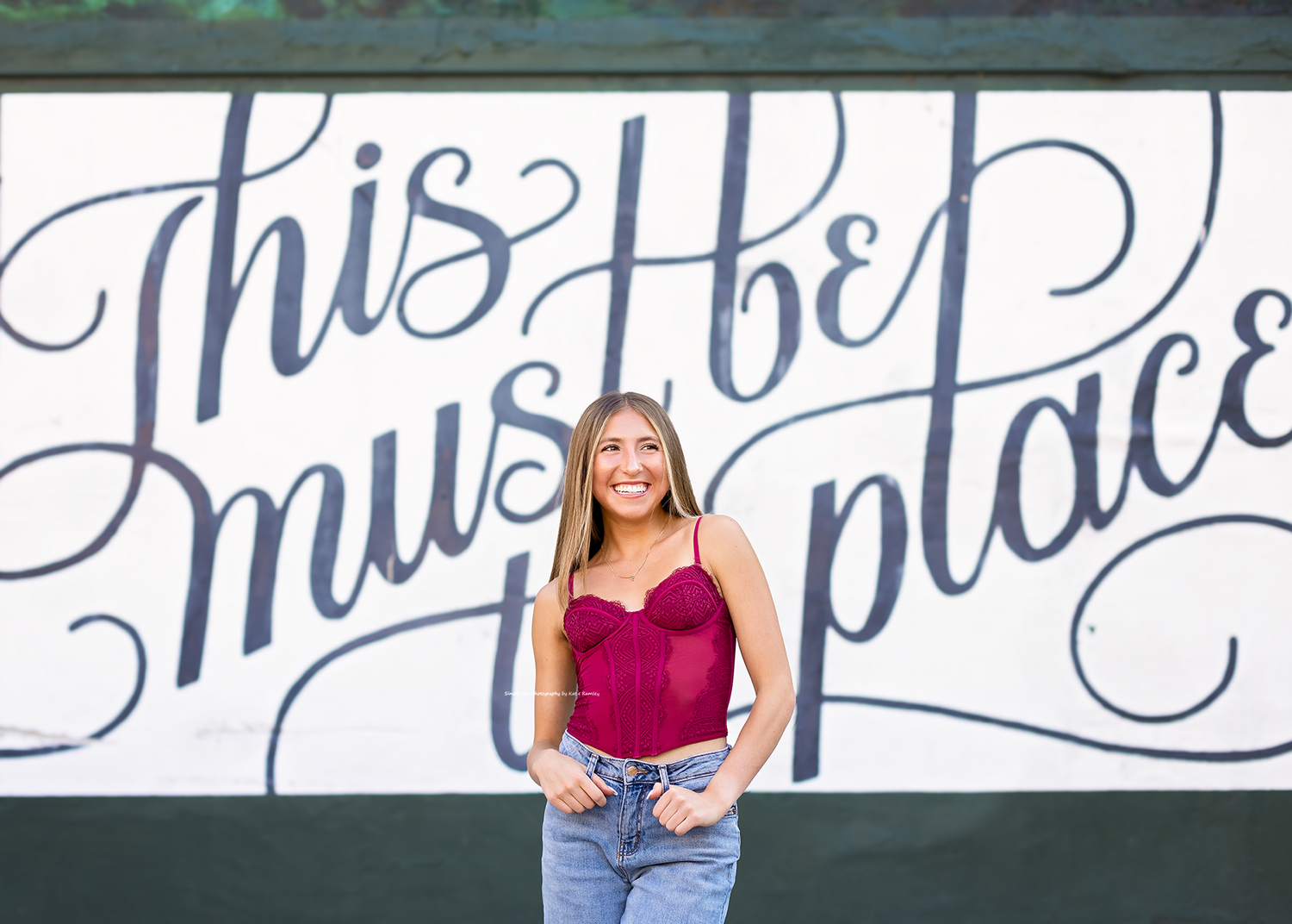 Young woman in red top and jeans smiling in front of a decorative mural reading This Must Be The Place.