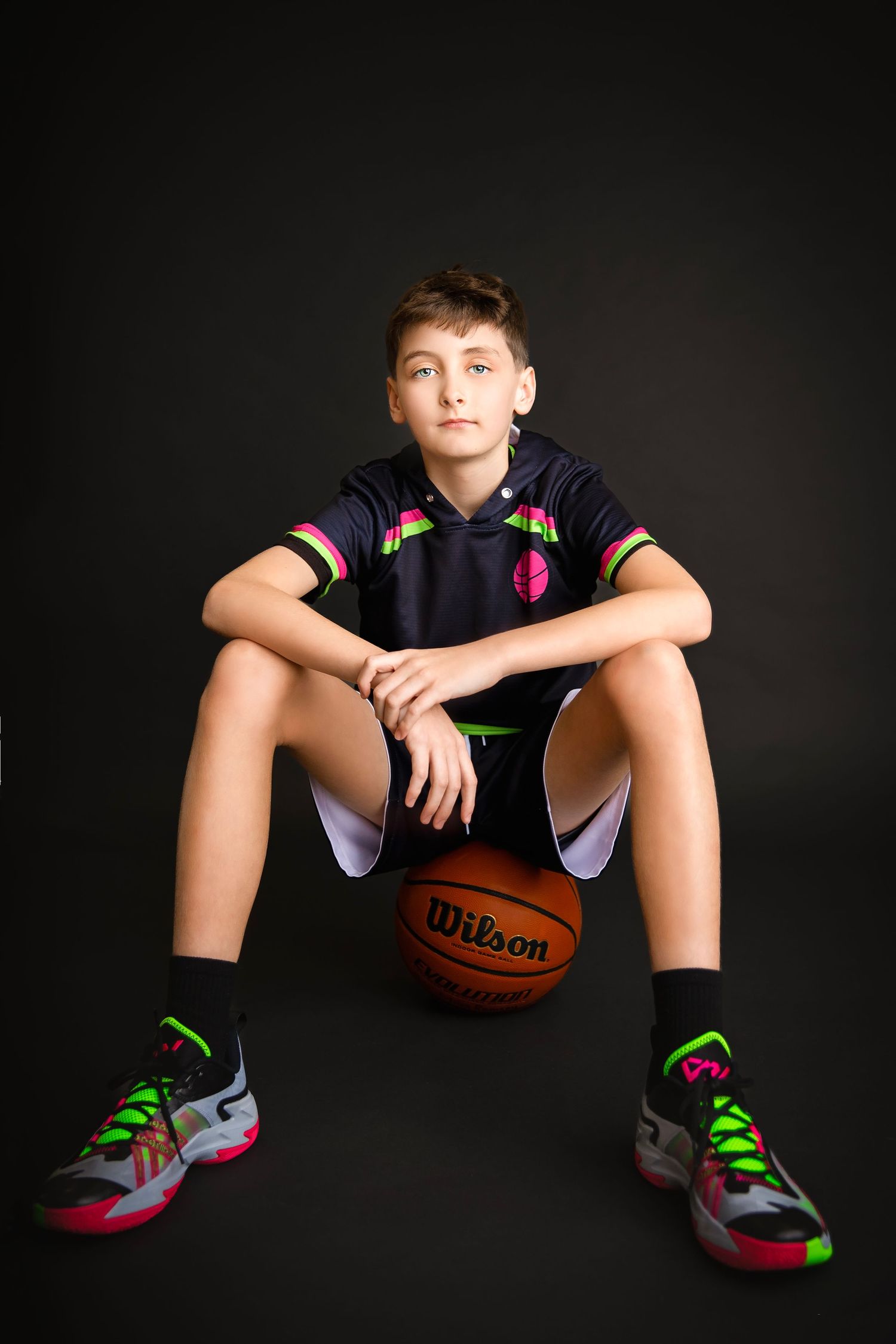 Young boy in black basketball uniform sitting on a Wilson basketball against a dark background.