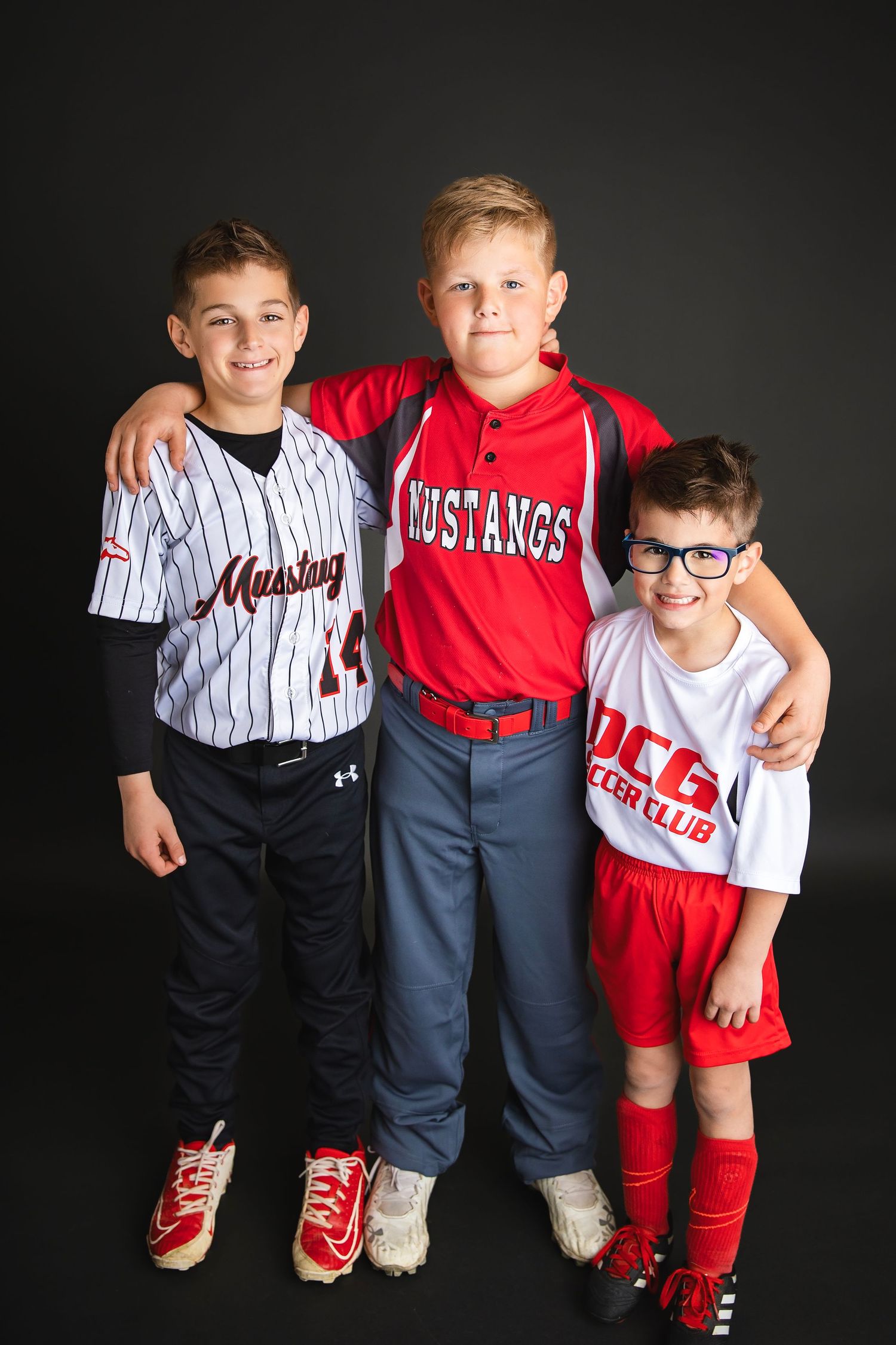 Three young brothers pose together in sports uniforms including two baseball jerseys and one soccer uniform.
