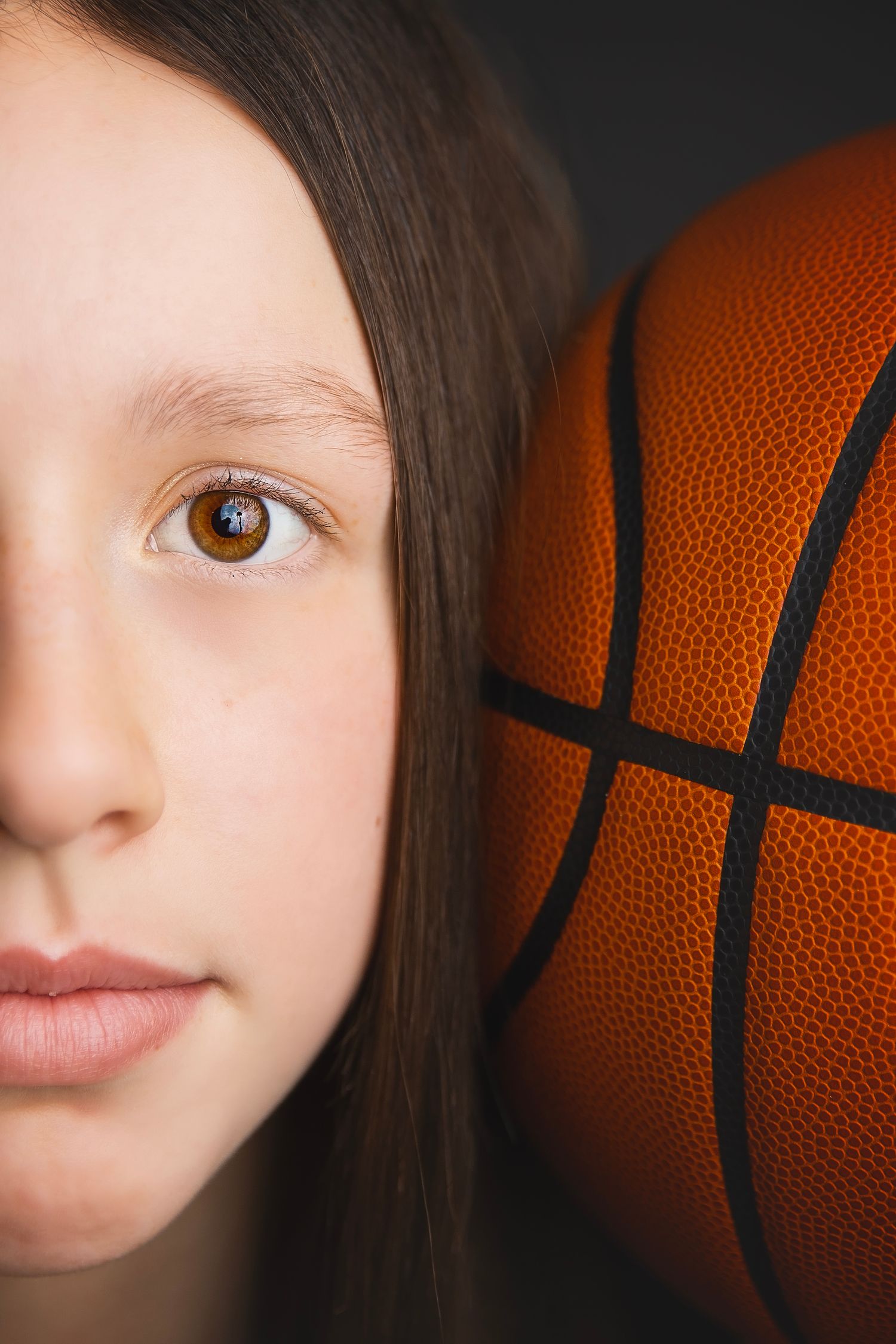 Close-up half portrait of a young girl beside an orange basketball on dark background.