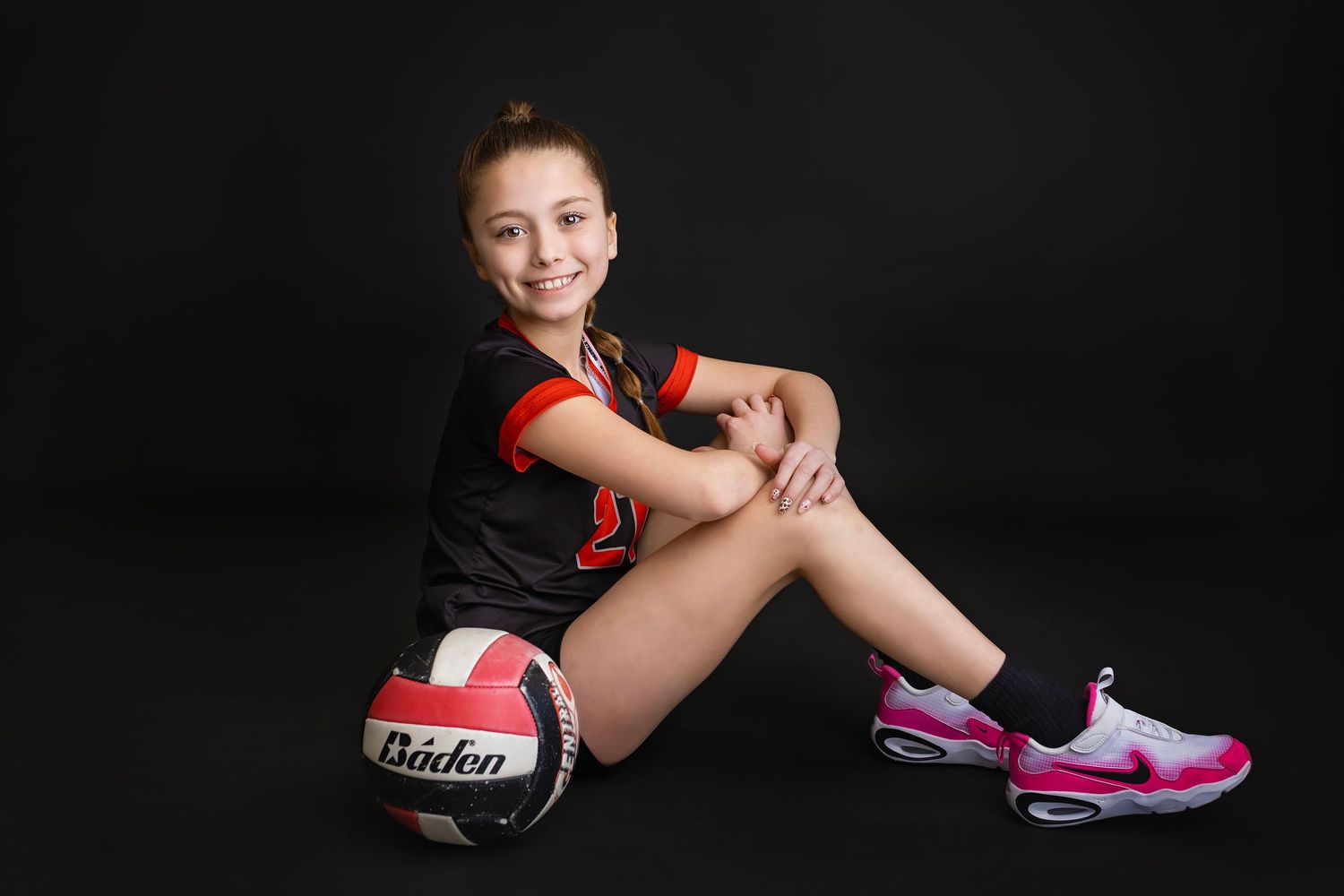 Young girl in black and red volleyball uniform sits smiling with a Baden volleyball beside her on a dark background.