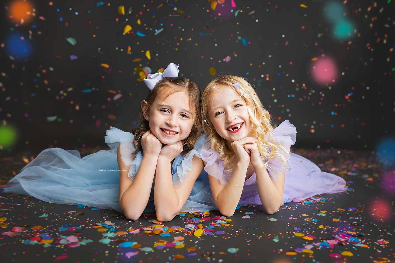 Two smiling girls in pastel tulle dresses lying on floor with colorful confetti.
