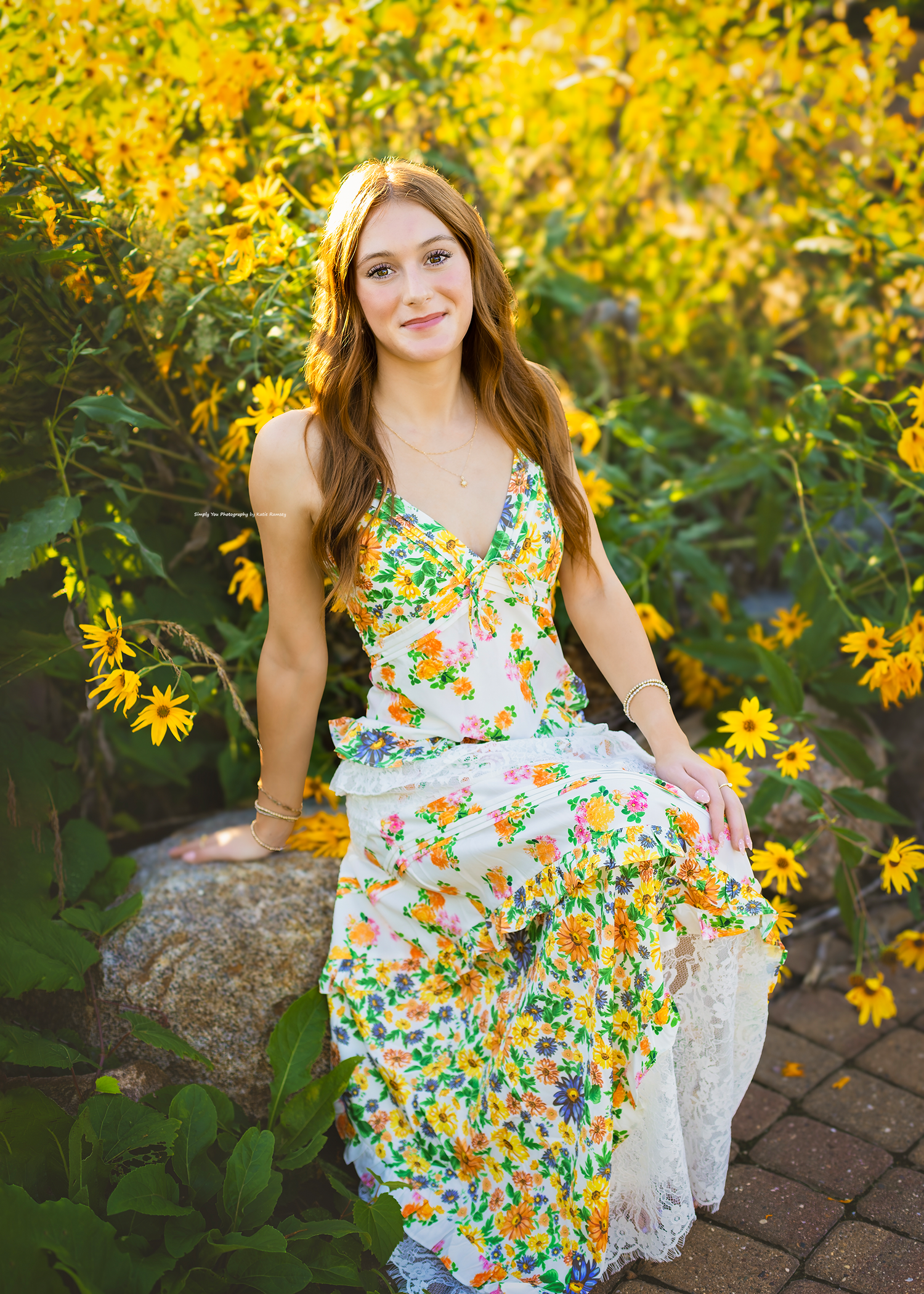 Young woman in floral maxi dress sitting on a rock surrounded by vibrant yellow wildflowers in a garden.