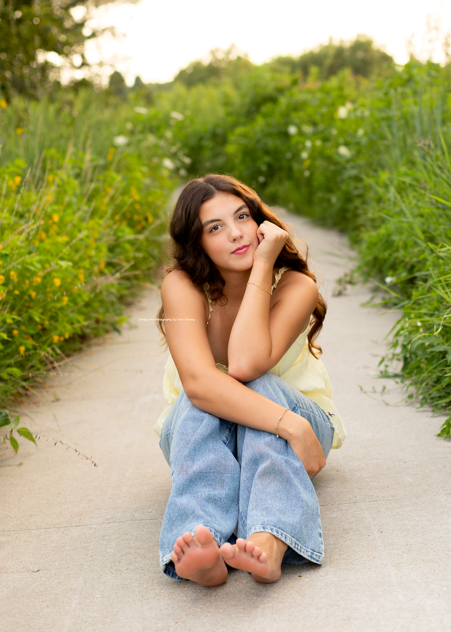 Young woman in jeans and yellow top sitting barefoot on a path surrounded by lush green meadow at golden hour.