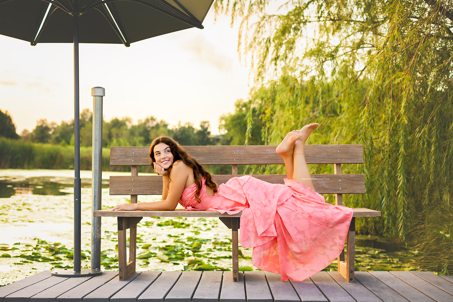 Woman in pink dress relaxing on bench by lily pond under umbrella near weeping willow at sunset.