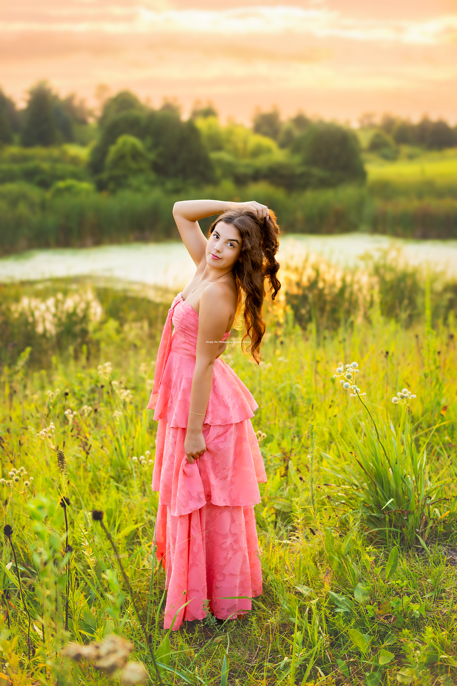 Young woman in pink ruffled dress standing in lush green meadow near lake at golden sunset.