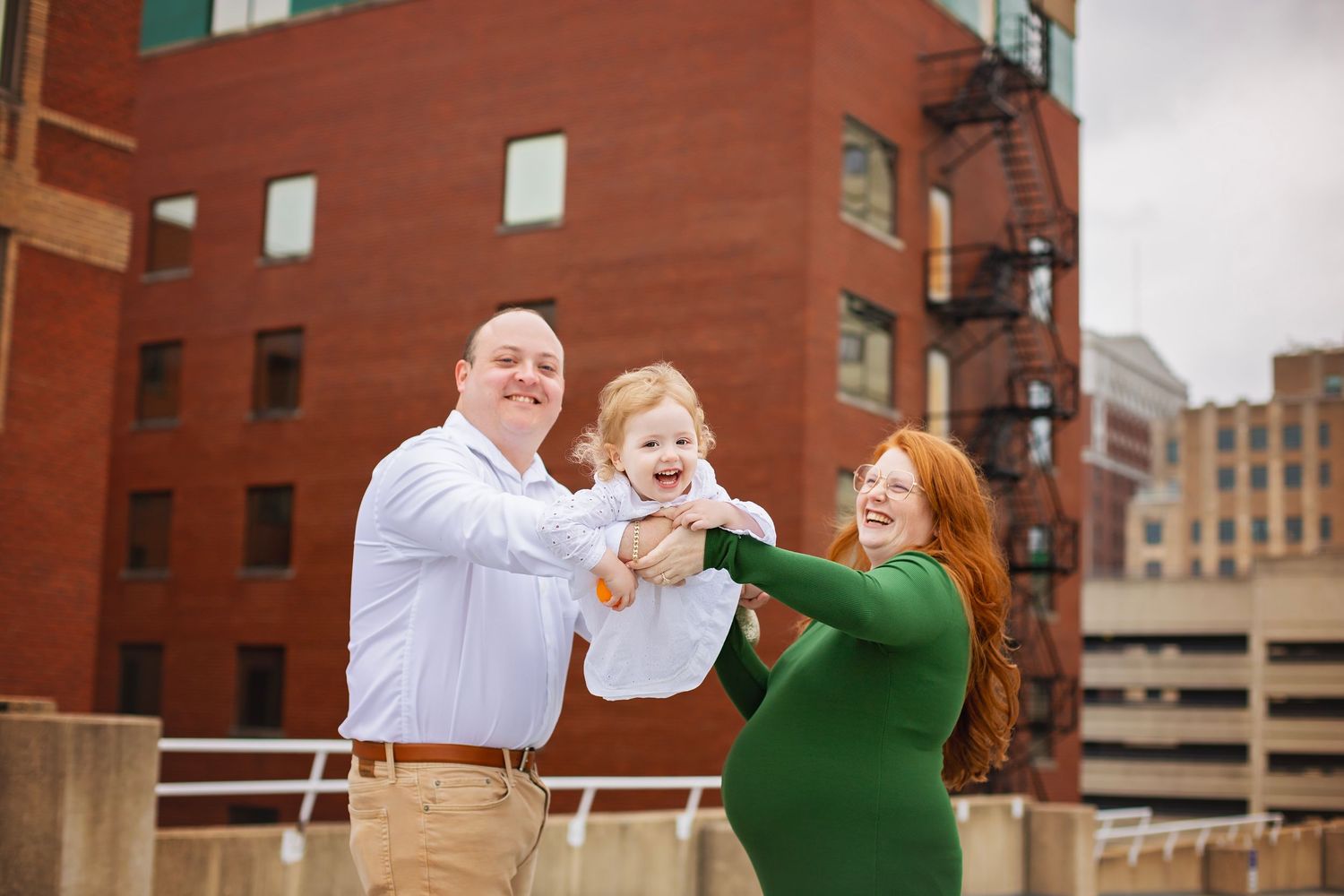 A joyful family lifts their laughing toddler outdoors near a brick building.