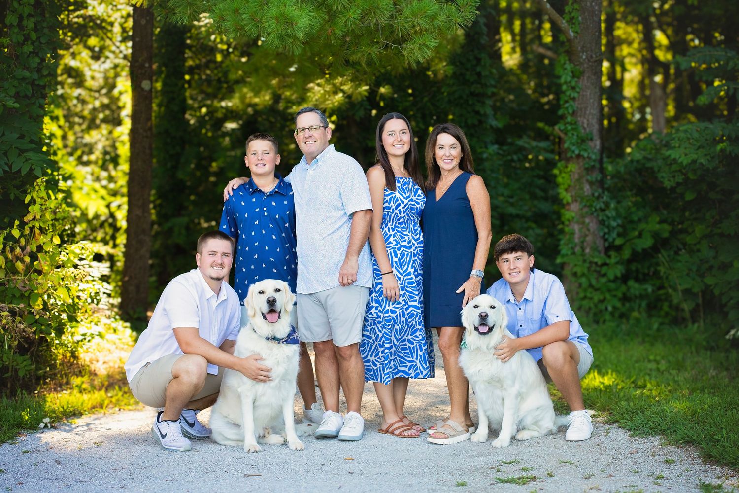 Family of six posing with two large white dogs on a shaded gravel path in a wooded area.