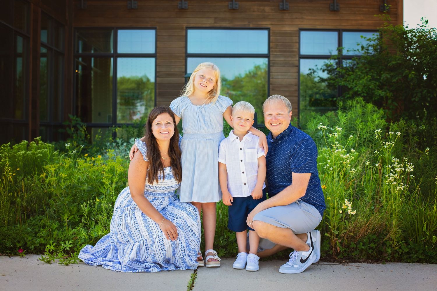 Family of four poses together outside a modern building surrounded by lush green gardens on a sunny day.
