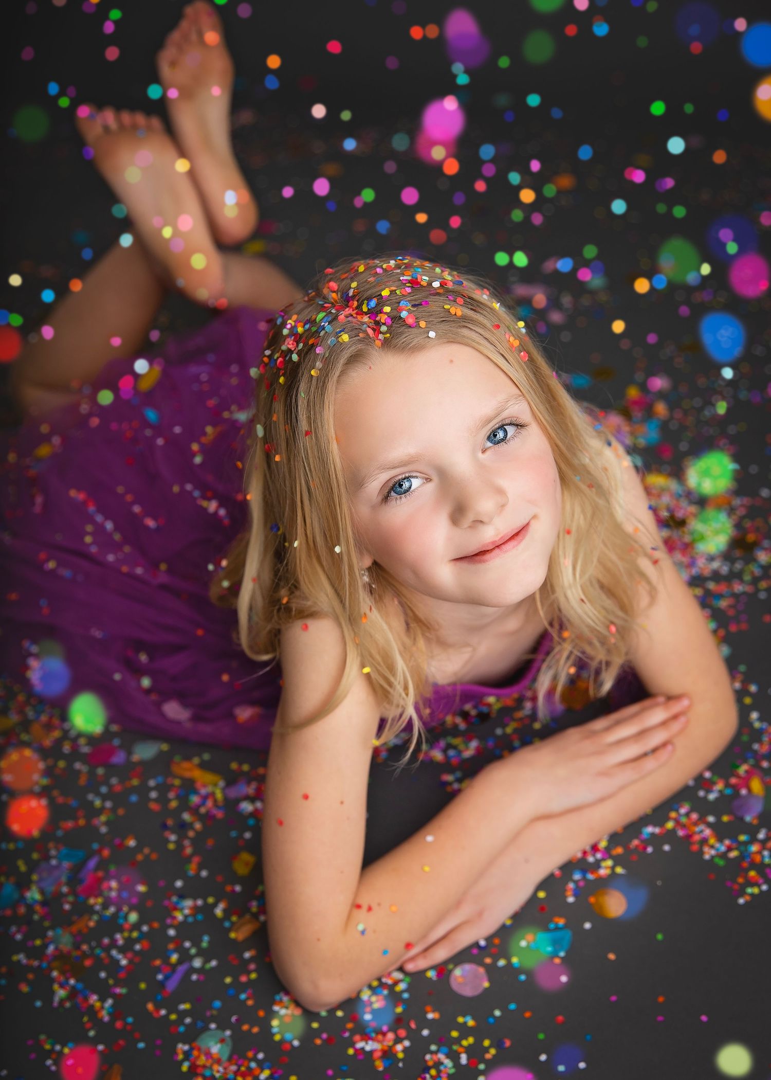 A smiling blonde girl in a purple dress lies on a floor covered in colorful confetti with bokeh light effects.