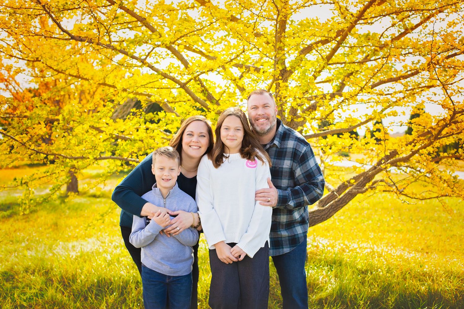 Family of four smiling in front of a bright yellow fall tree.