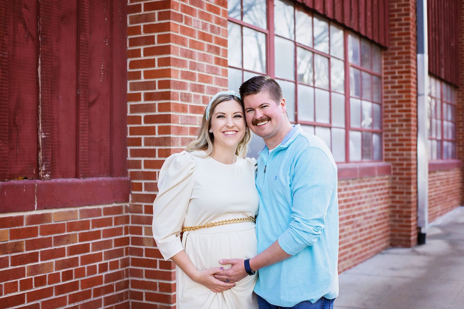 Smiling couple posing together during maternity photo session by red brick wall.