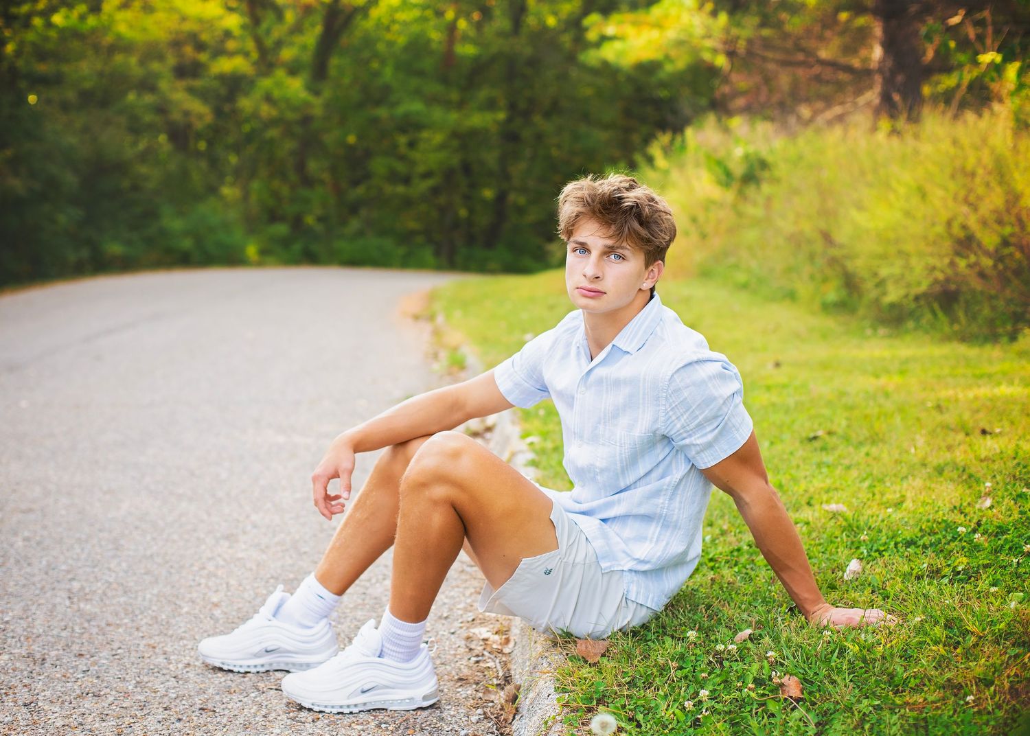 Teen boy in light blue shirt and shorts sits on a curb beside a tree-lined road, wearing white sneakers.