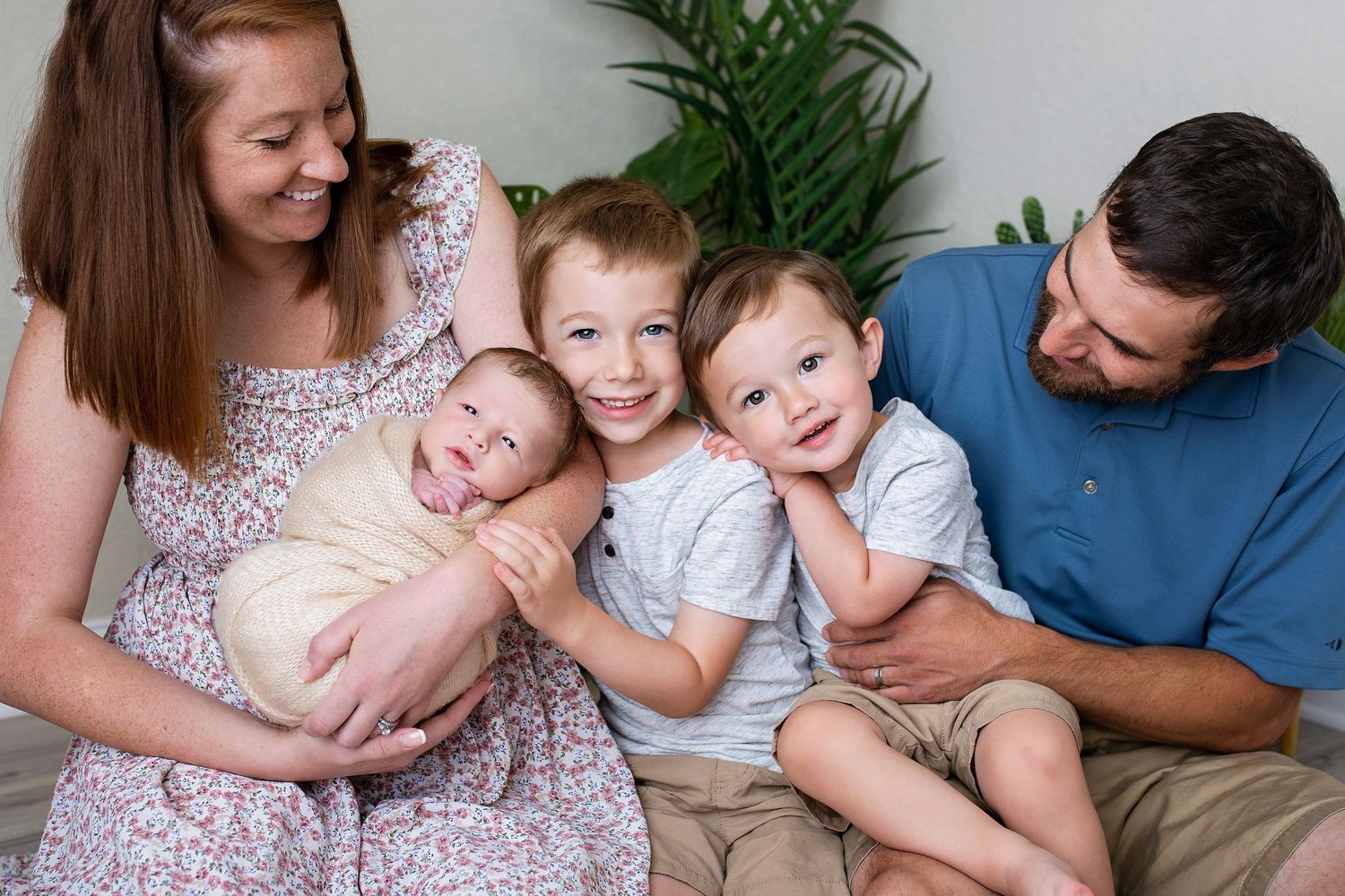 Happy family of five with newborn baby, two young boys, mother and father sitting together smiling.