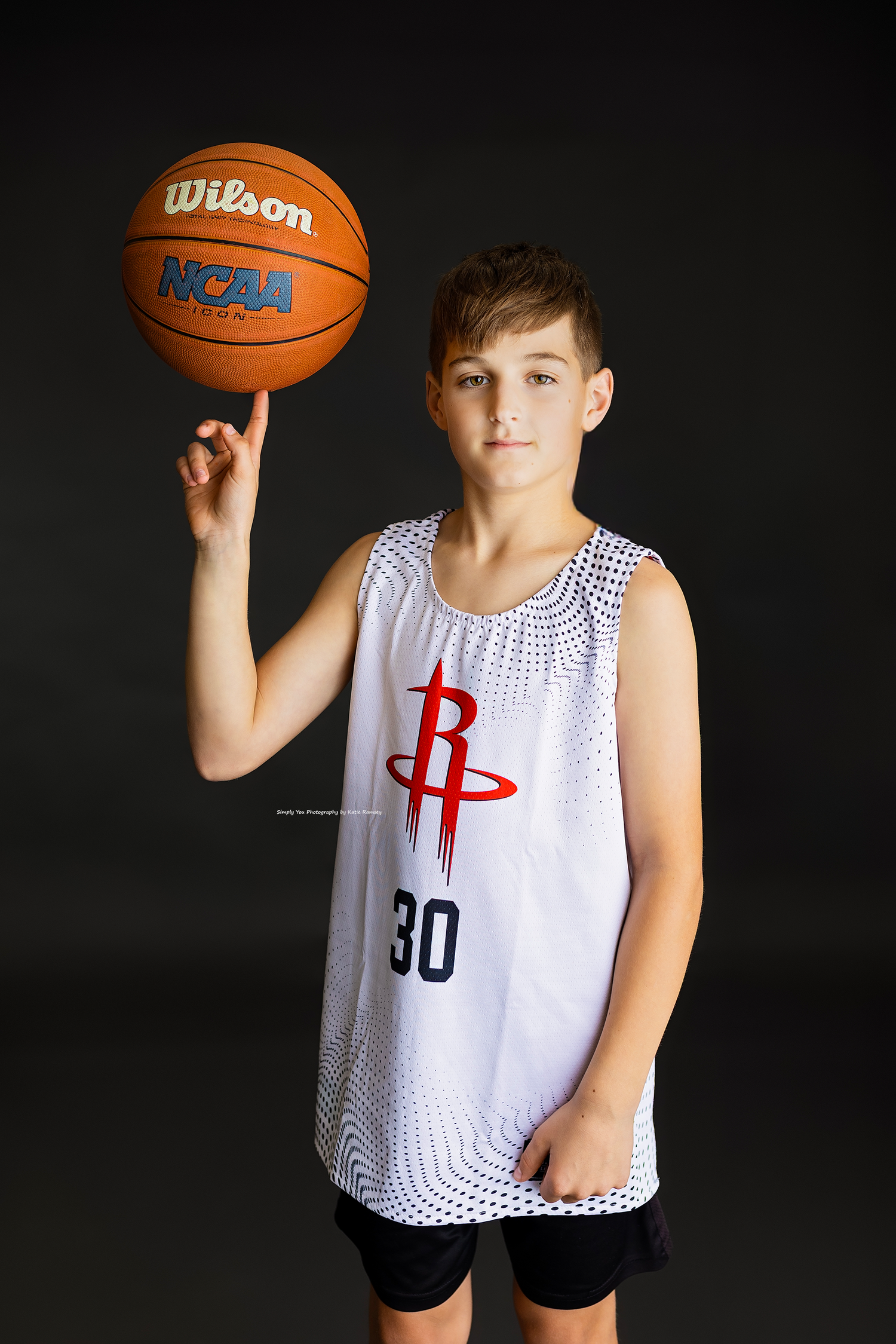 Young boy in Houston Rockets jersey number 30 spins Wilson NCAA basketball on finger.