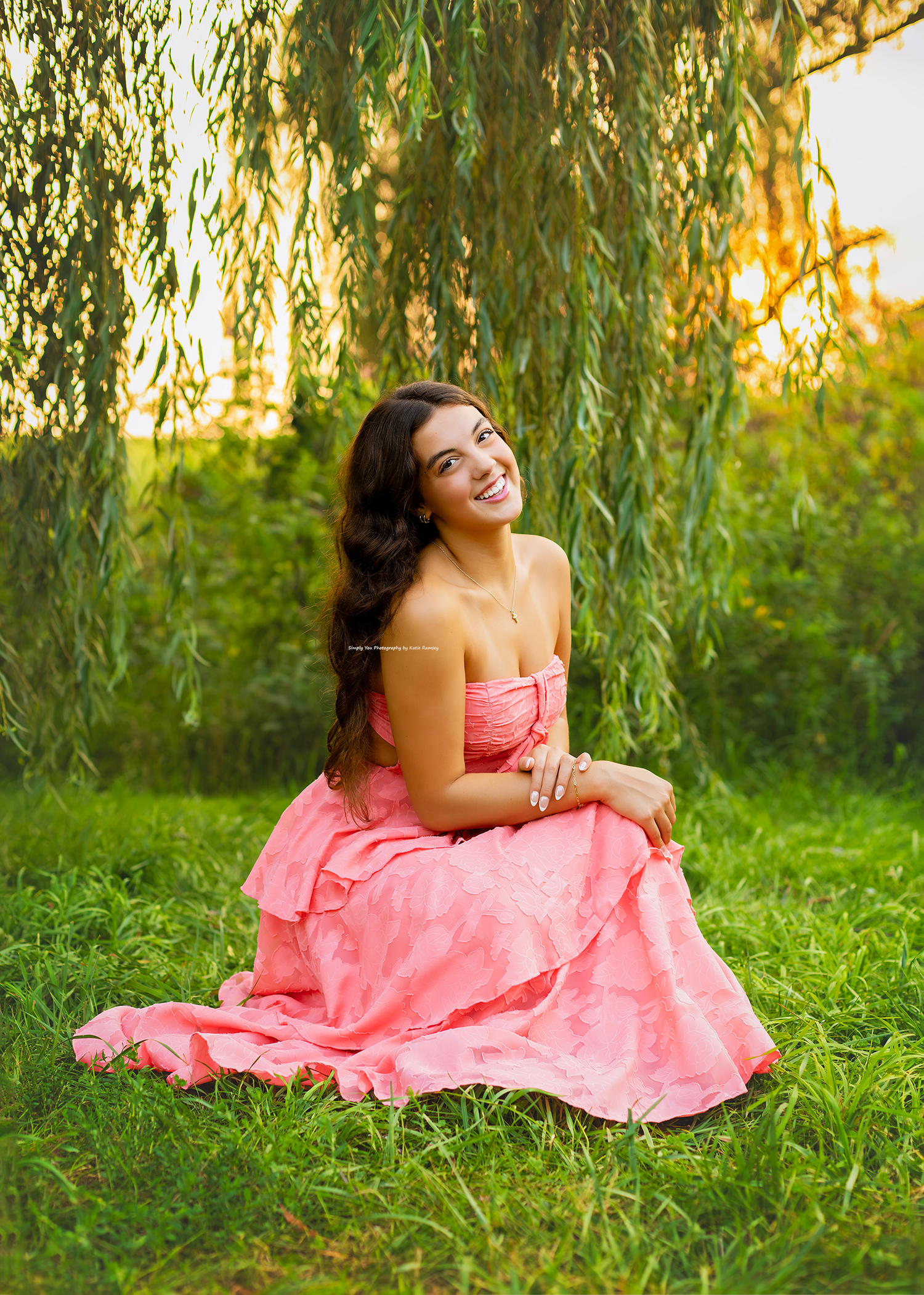 A young woman in a pink strapless gown sits gracefully on green grass beneath a weeping willow at golden hour.