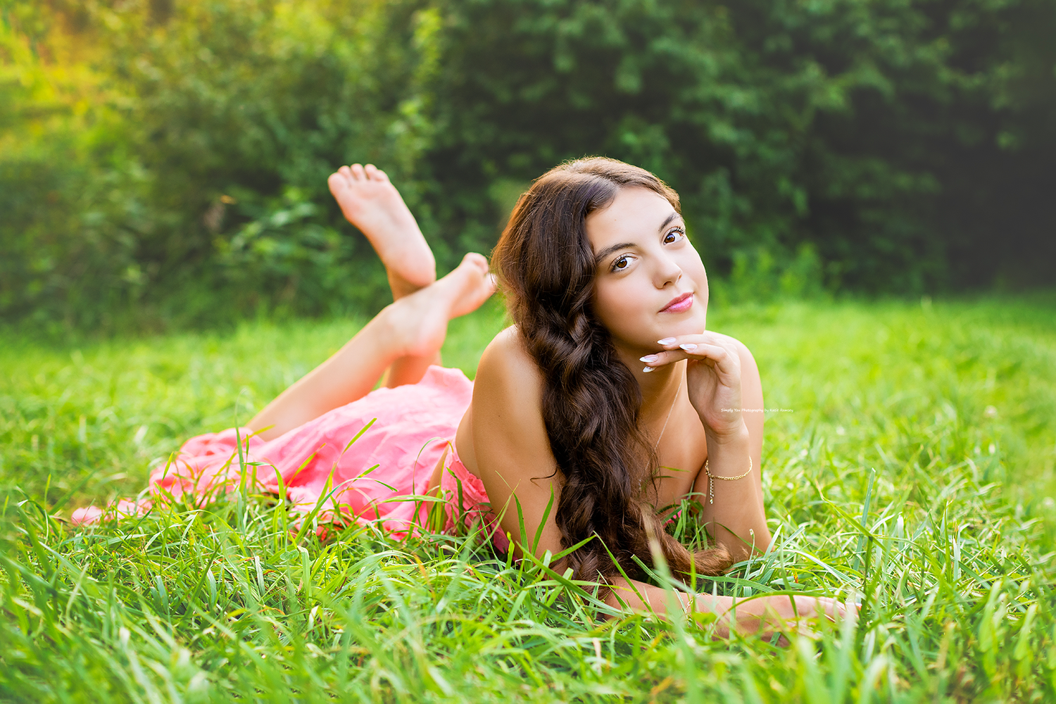 Young woman in pink dress lying on lush green grass, smiling peacefully in a sunny outdoor setting.