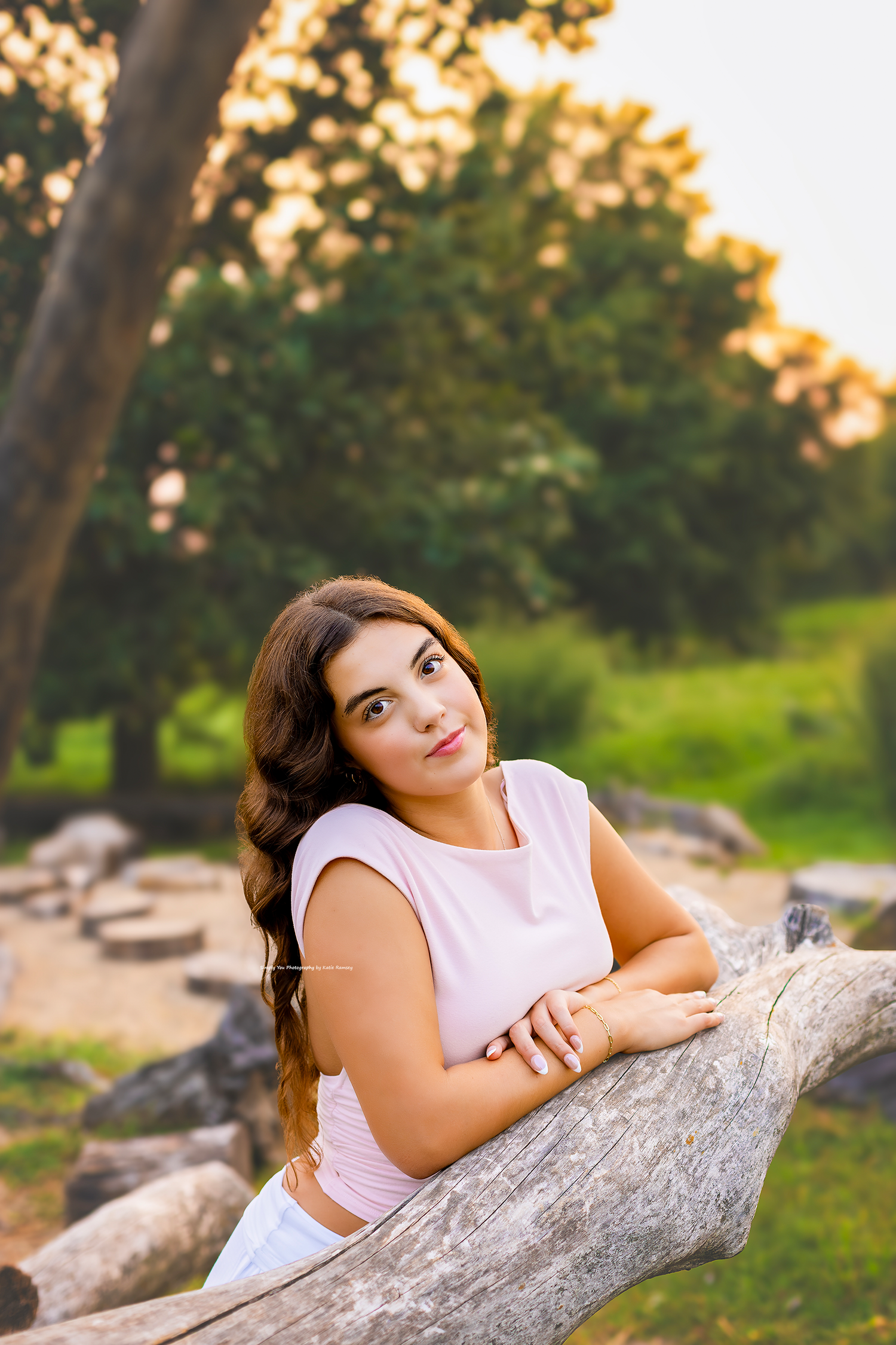 Young woman in pink top leaning on a wooden log in a sunlit park, looking up thoughtfully.