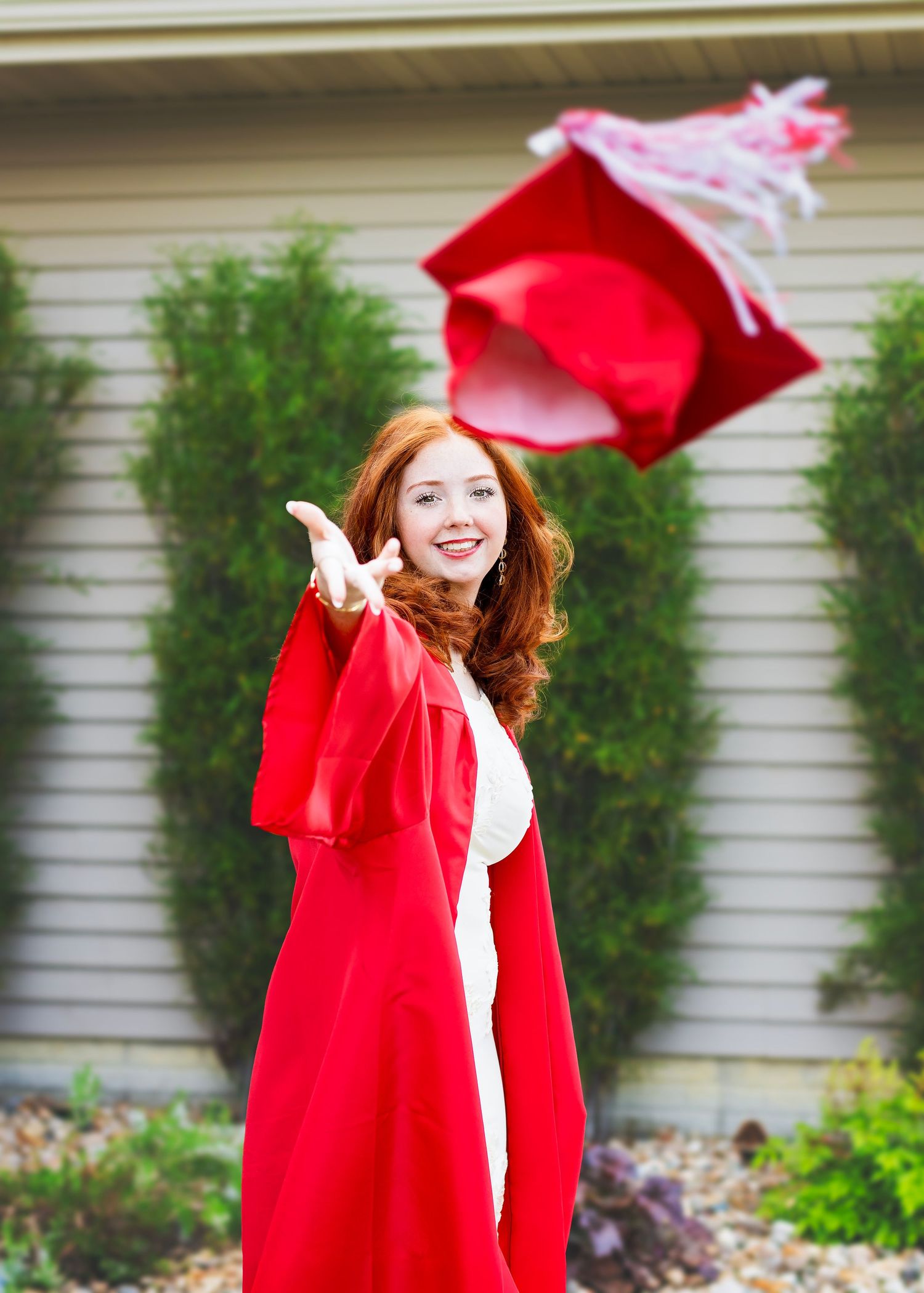 Smiling redhead graduate in red gown tossing cap in air during outdoor senior photo session.
