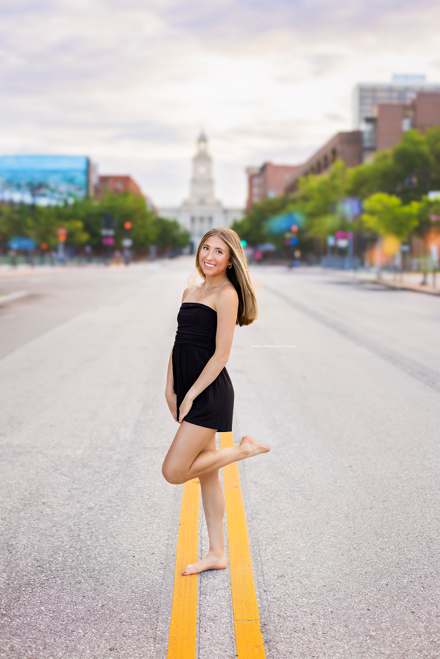 Young woman in black strapless dress posing barefoot on yellow road lines with city buildings in background.