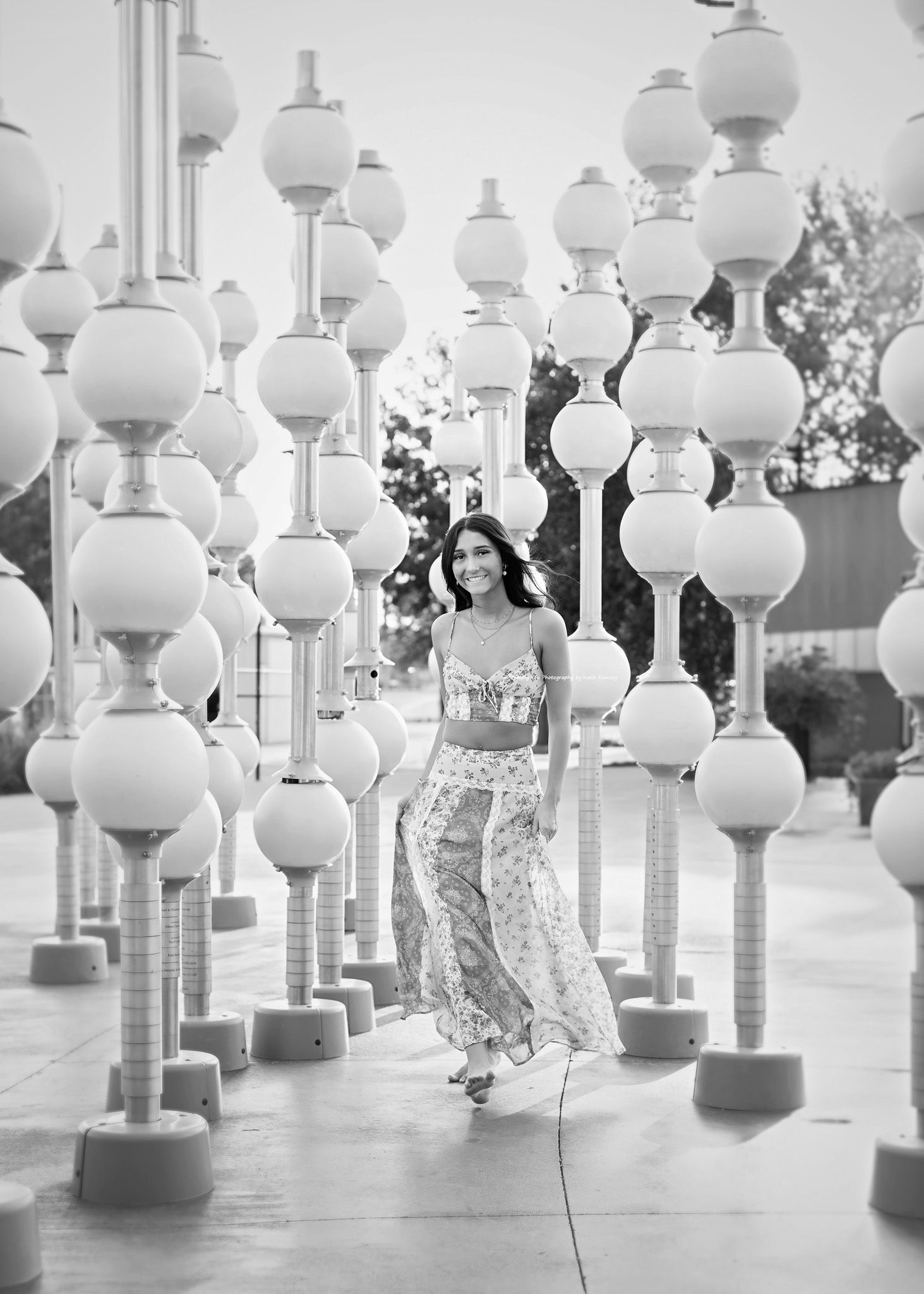 Black and white photo of a woman in floral skirt walking among spherical light installations outdoors.