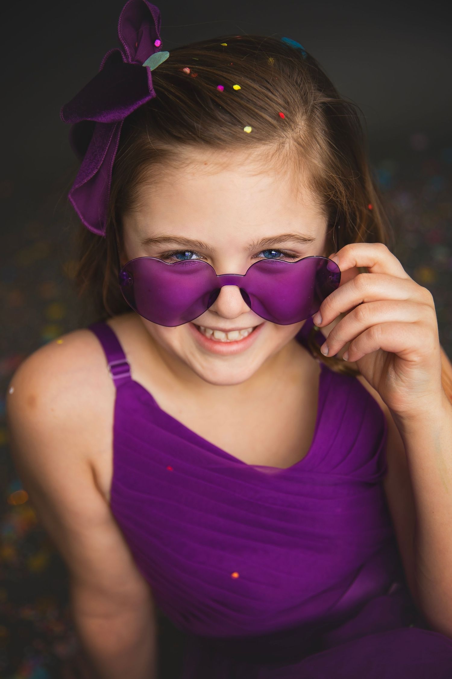 Playful girl in purple dress peering over heart-shaped purple sunglasses with a mischievous smile.