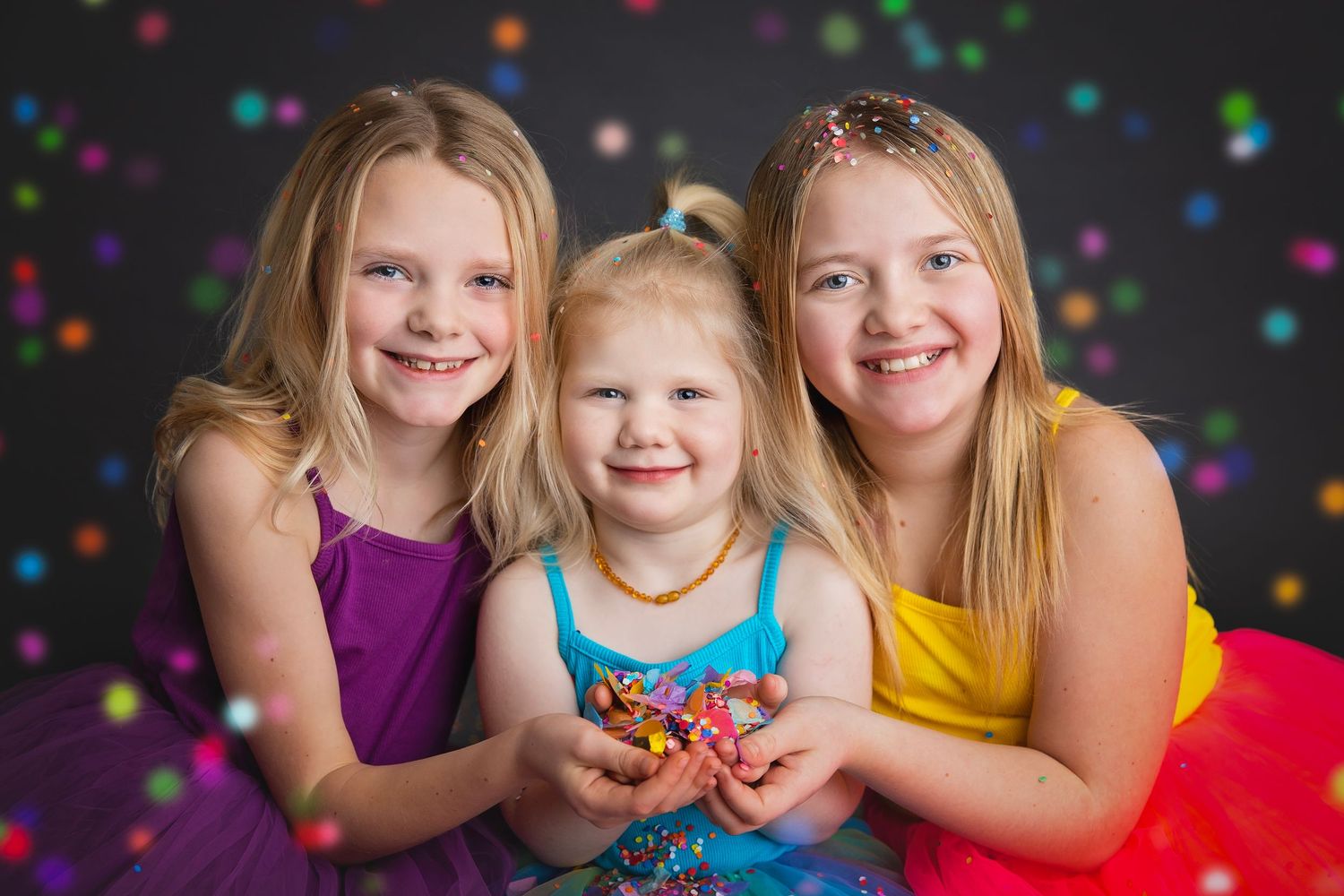 Three smiling blonde girls in colorful dresses pose together with confetti and bokeh lights in a festive studio photo.