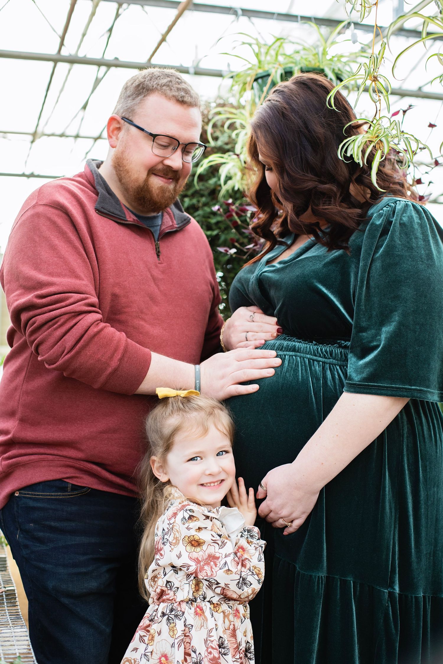 Father and young daughter touch pregnant mothers belly in a warm greenhouse maternity photo session.