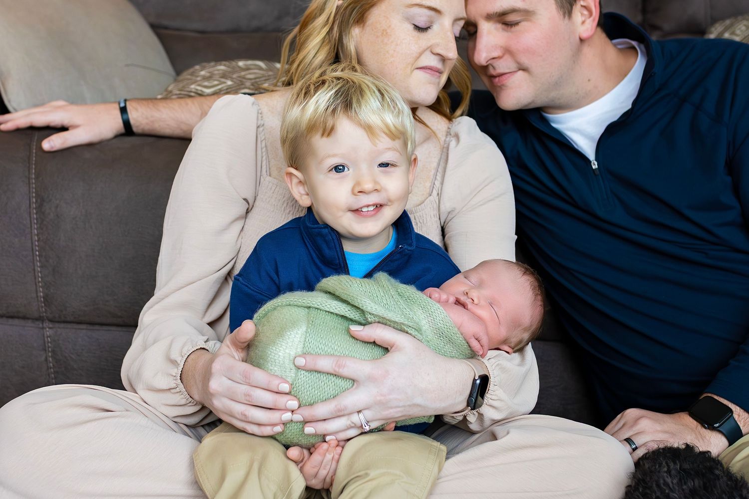 Young boy proudly holds swaddled newborn sibling while parents smile lovingly on couch.