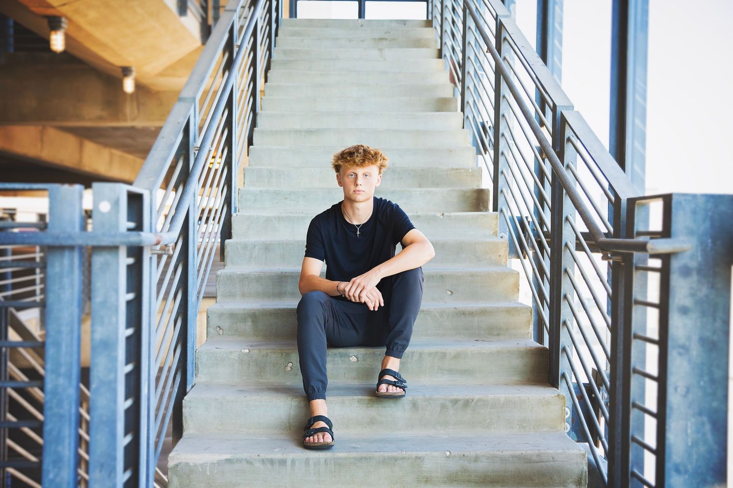 Teen boy with curly red hair sits on concrete stairs with metal railings in a modern building.