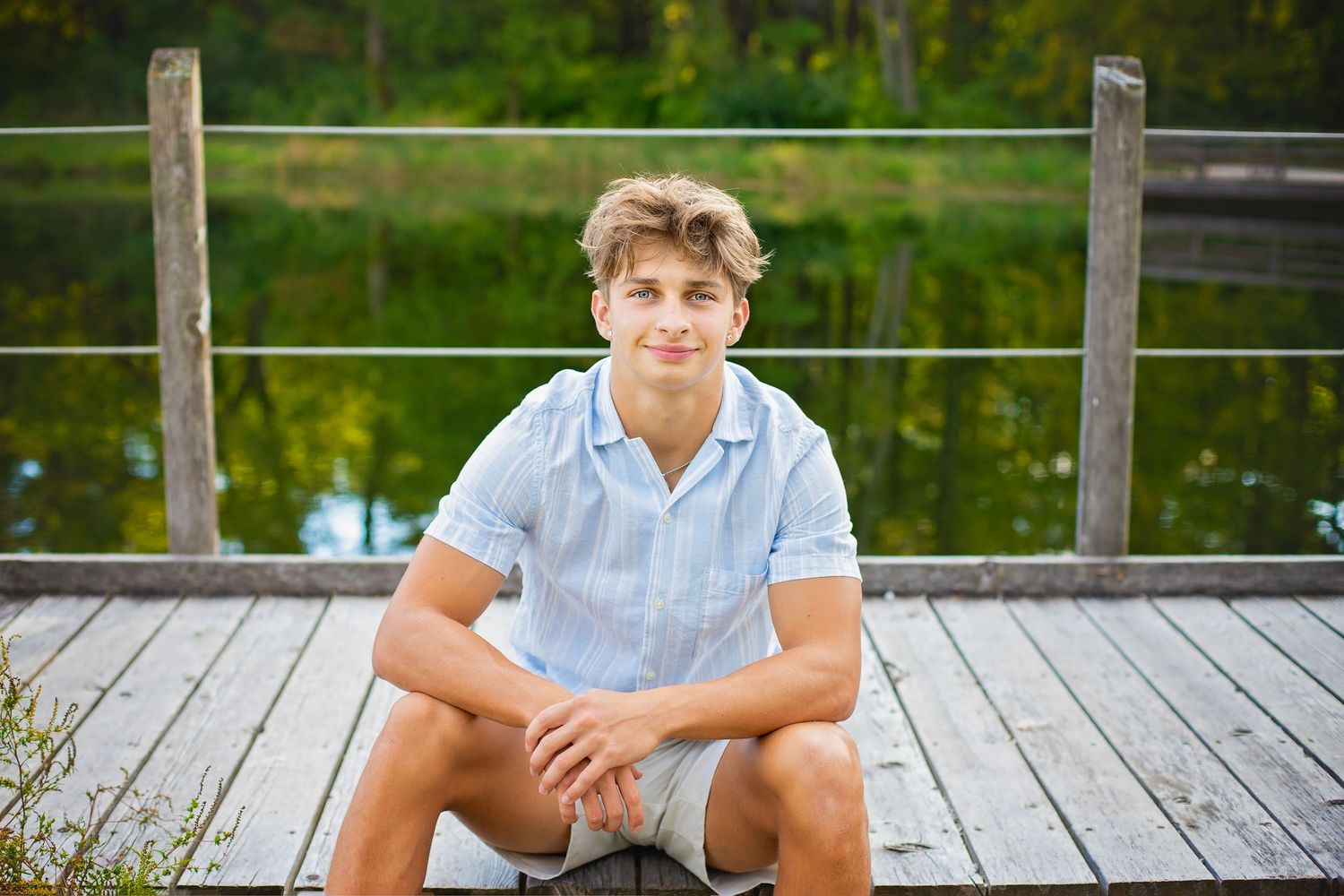 Blond teen boy in light blue shirt sits on a wooden dock near a pond with green trees behind him.
