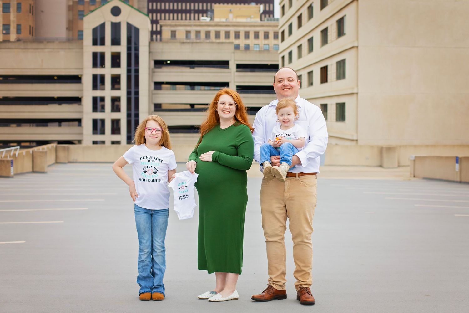 Family holding baby onesies and shoes for maternity announcement on urban rooftop parking garage.